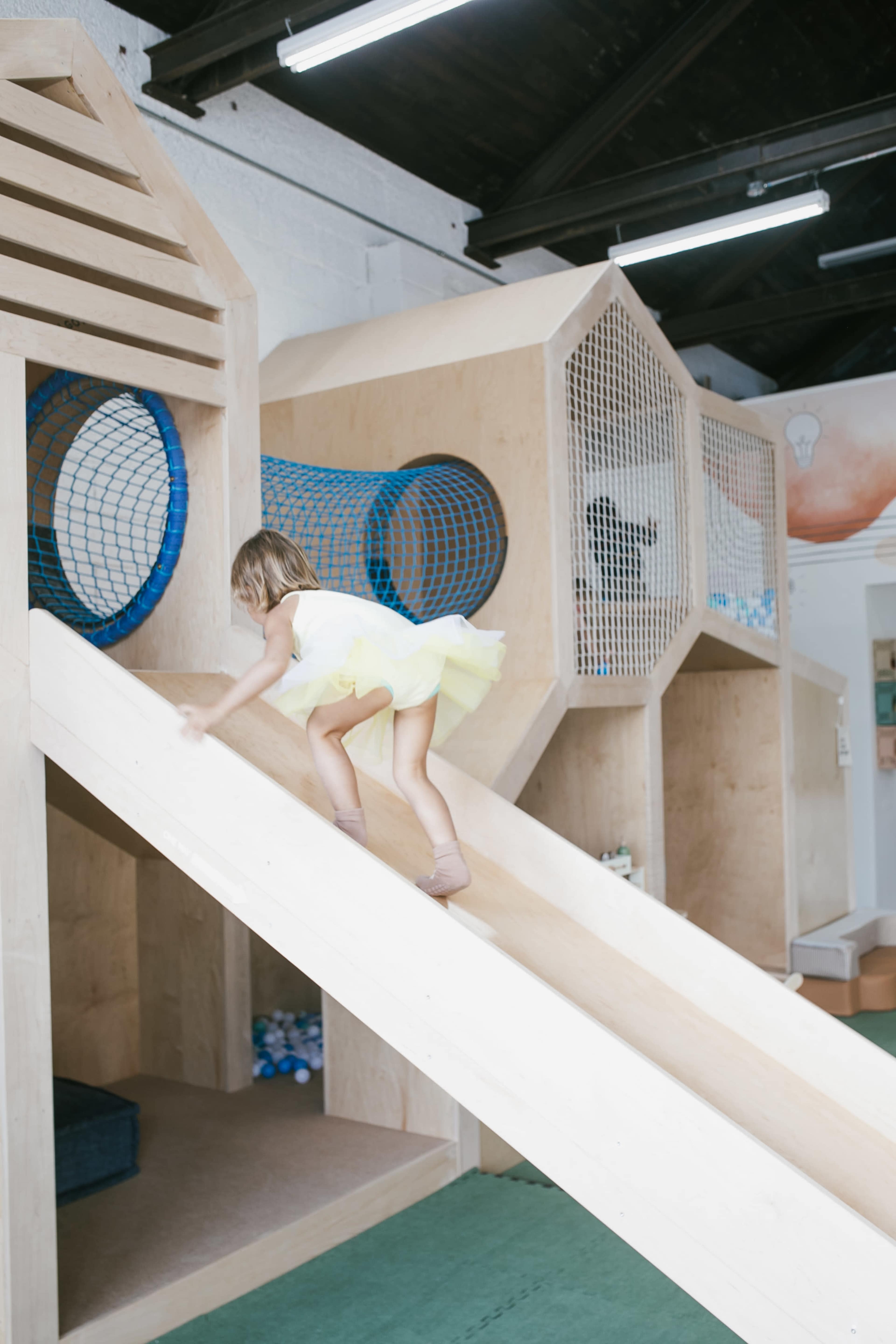 A child in a yellow dress climbs a wooden slide in a play area designed with geometric structures.
