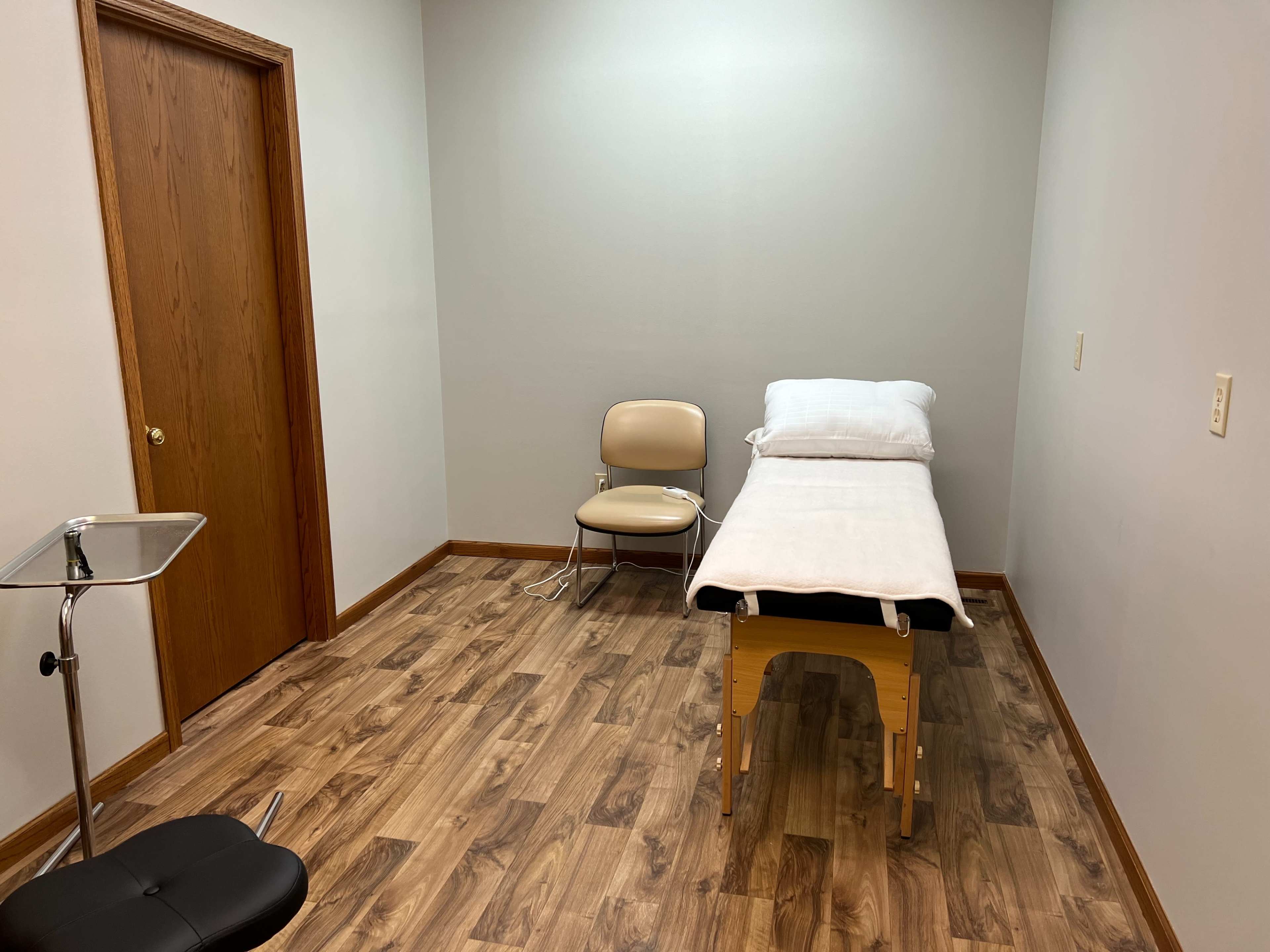 A plain examination room with a massage table covered by a white sheet, a chair, and a small side table.