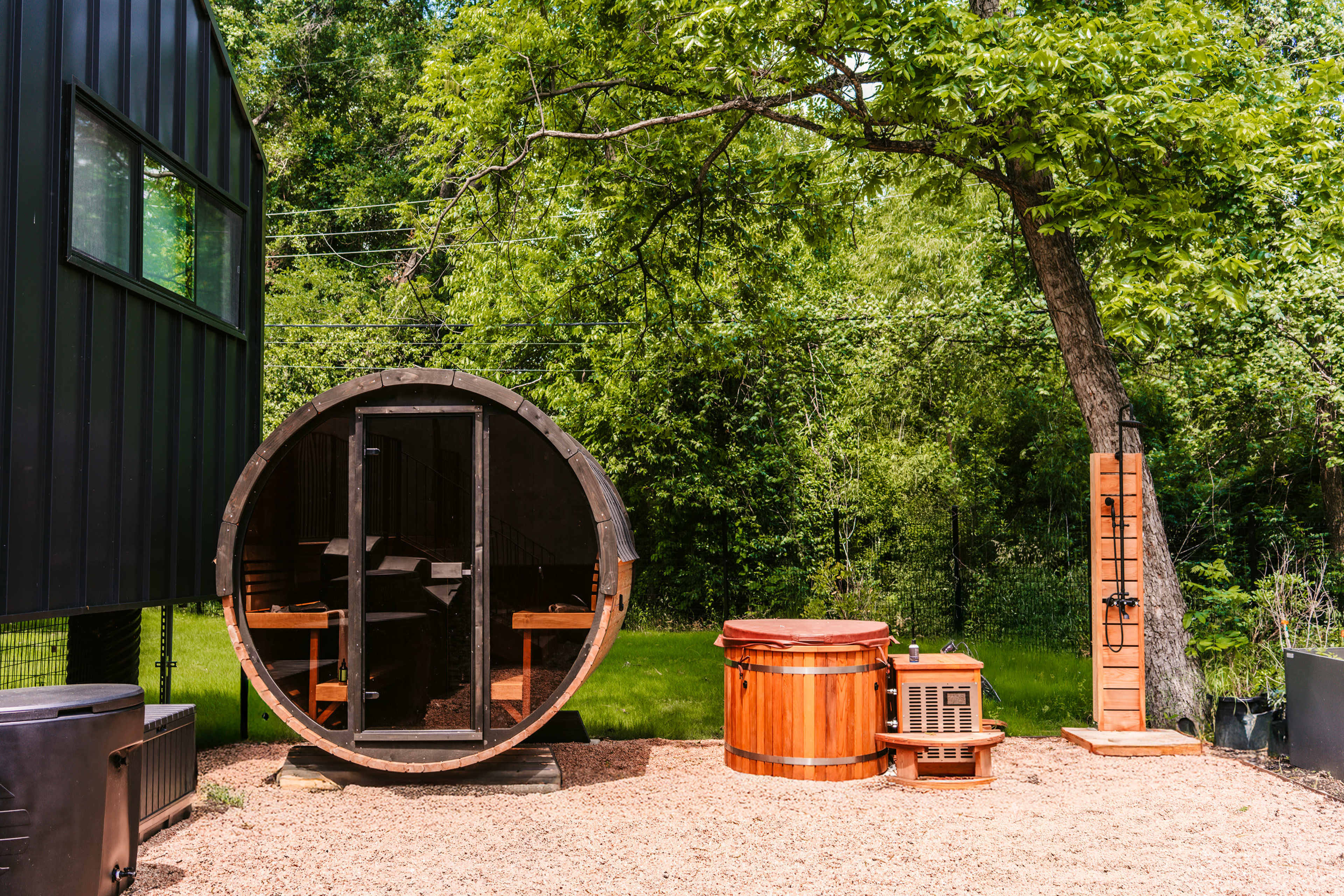 The image shows a circular sauna made of wood, a wooden hot tub, and an outdoor shower area, all situated in a green, grassy environment next to a black structure.
