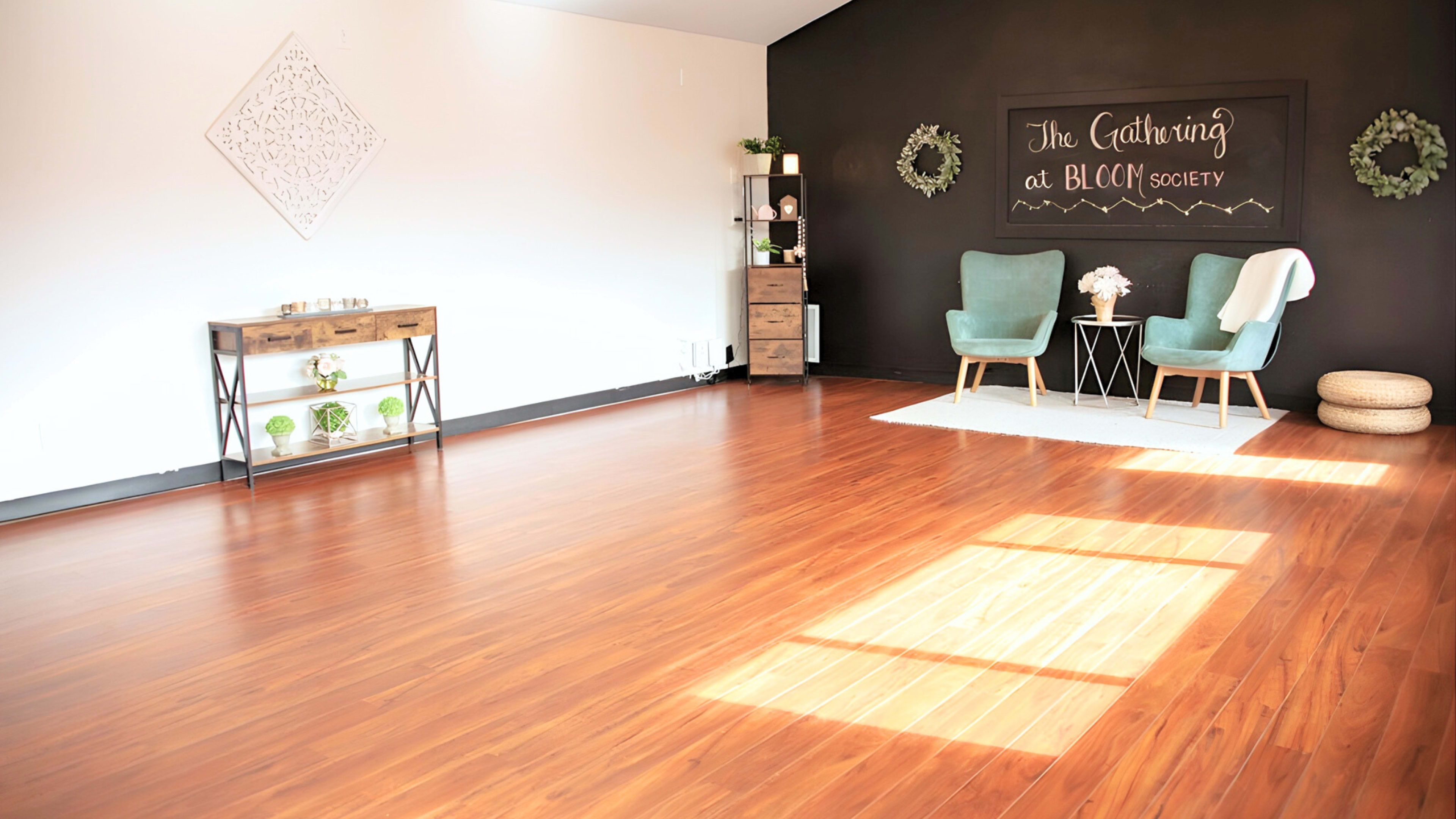 The image shows a spacious, well-lit room with wooden flooring, featuring two light blue chairs, a small black table, and a decorative wall with the text "The Gathering at BLOC Society."