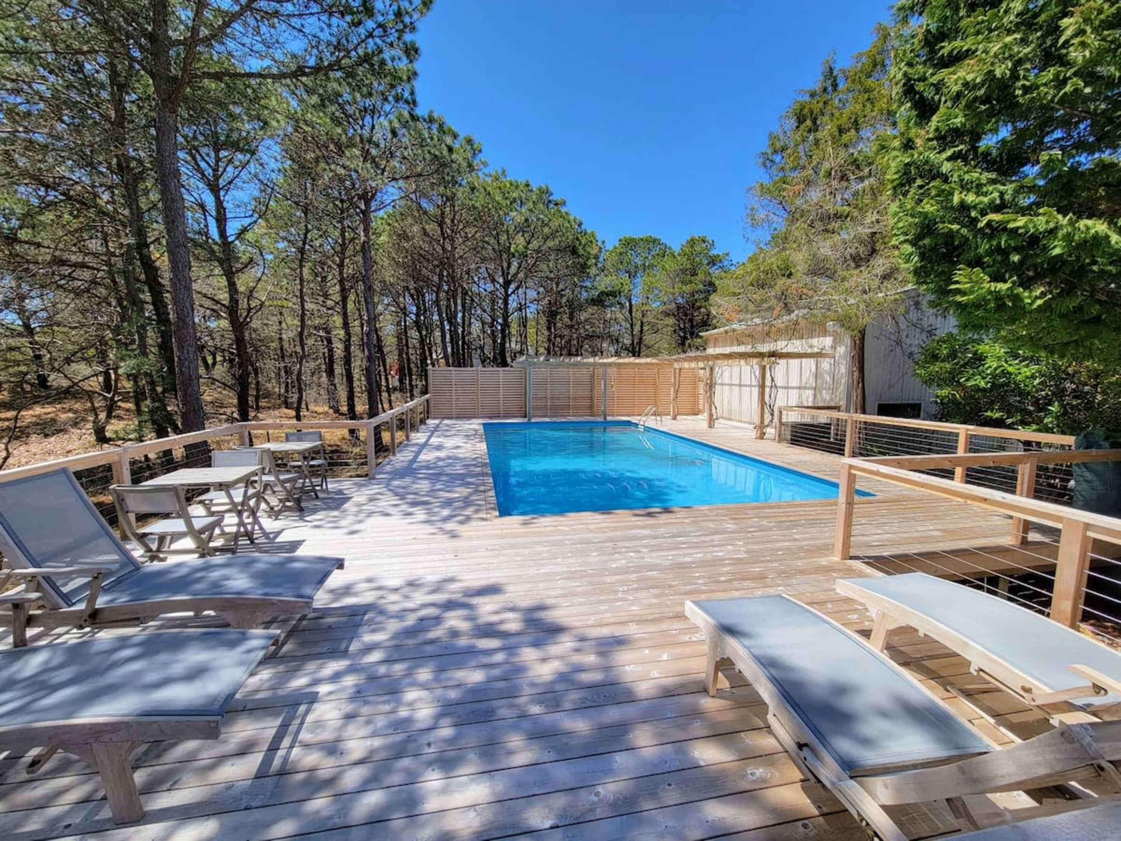 A rectangular pool surrounded by a wooden deck, with lounge chairs positioned nearby and trees in the background.