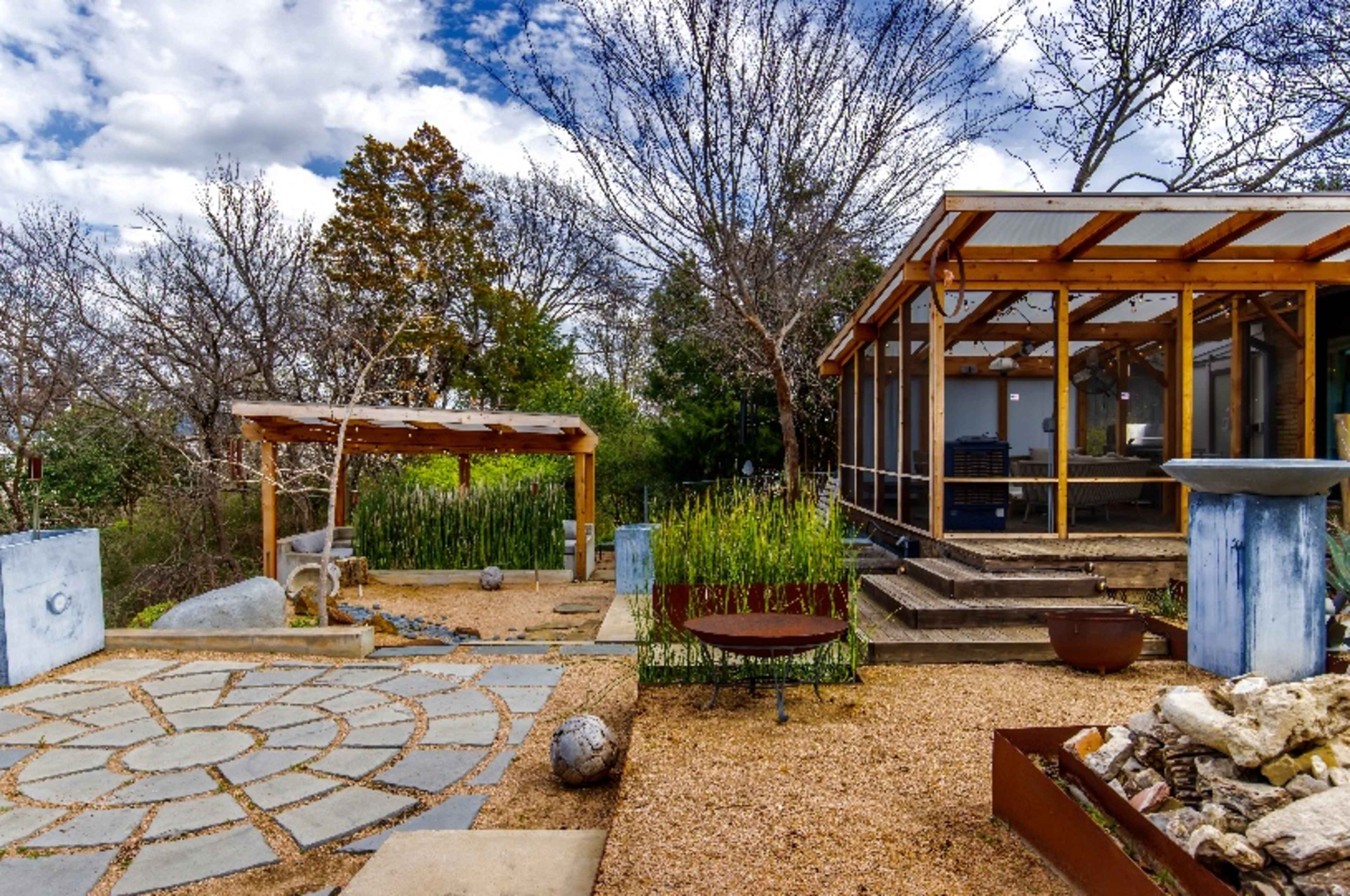 The image shows a landscaped outdoor area featuring a stone pathway, a wooden pergola, a screened-in structure, and a gravel section with various decorative elements.