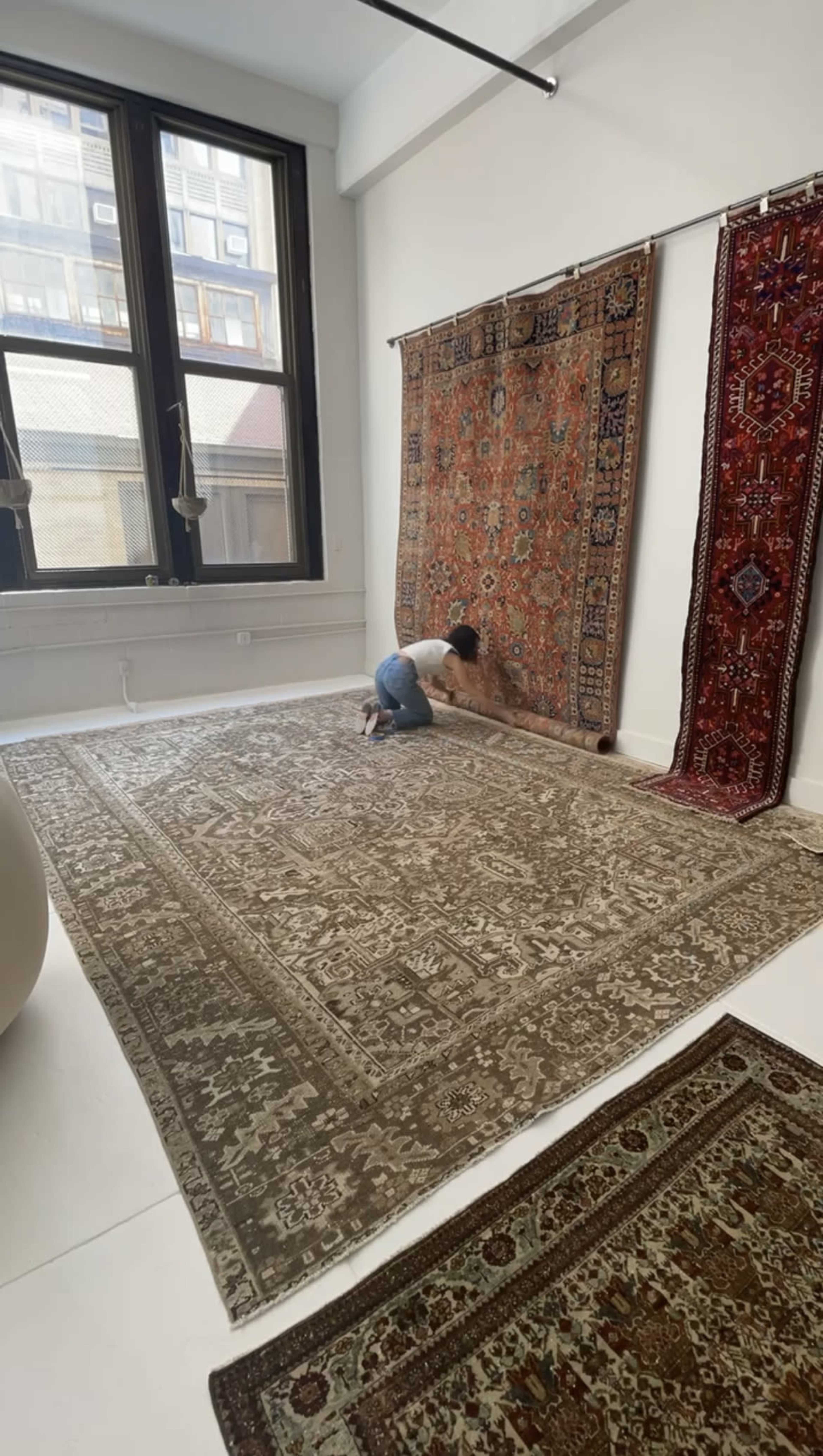 A person is kneeling on a patterned carpet in a brightly lit room with large windows while inspecting a hanging rug on the wall.