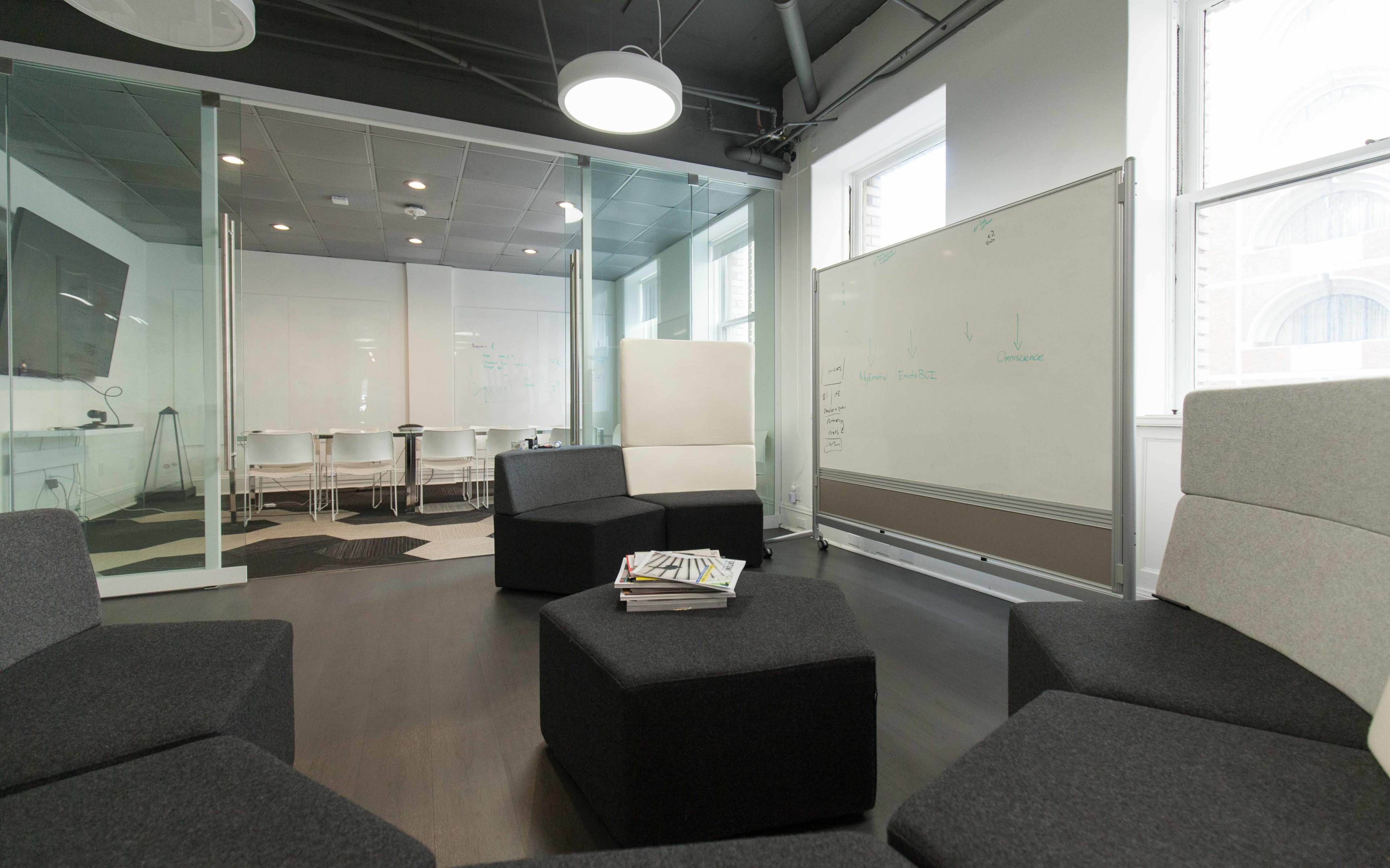 A modern office space featuring a seating area with modular black and gray ottomans, a whiteboard, and a glass-walled conference room with white chairs.