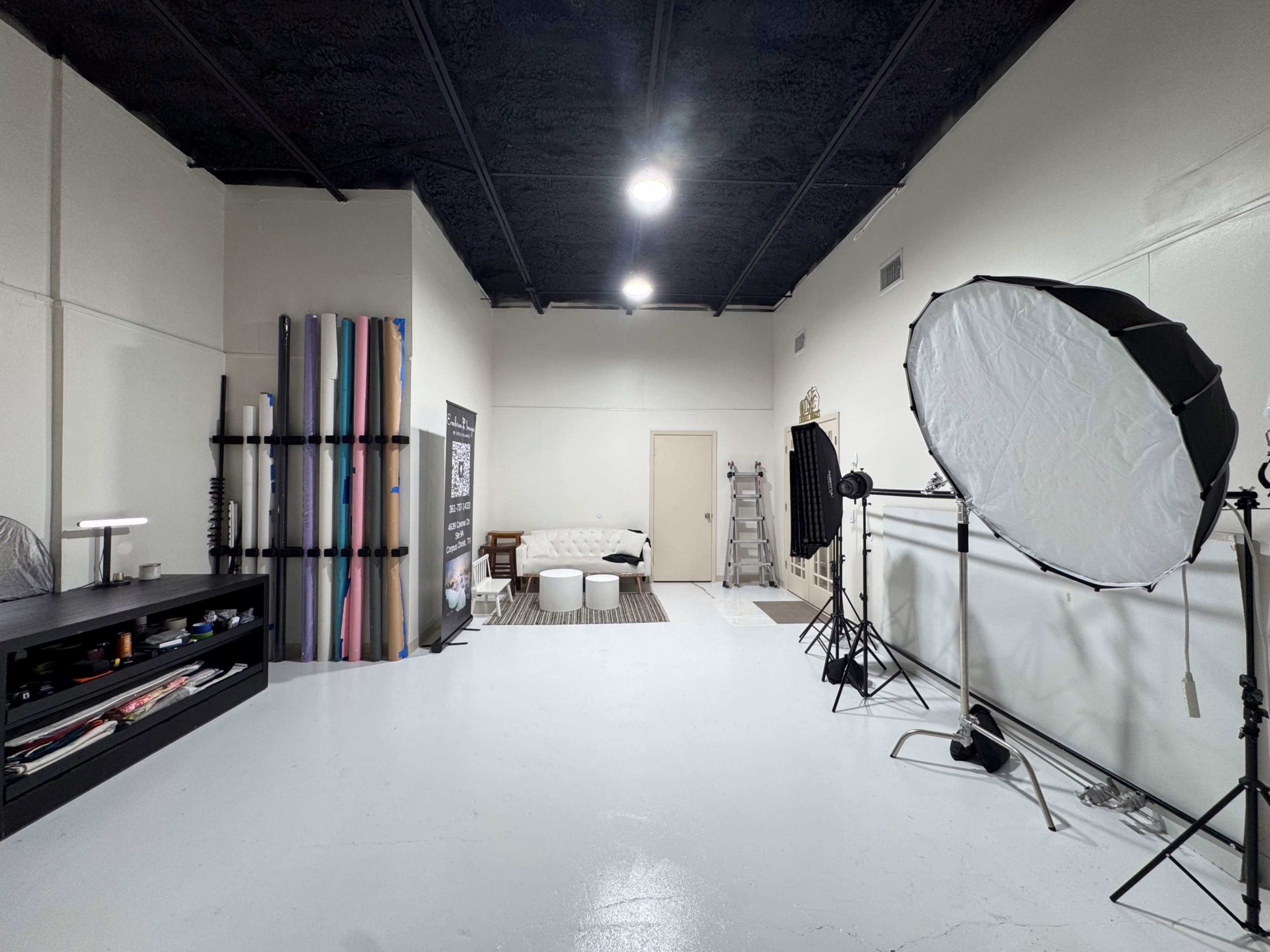 The image shows a photography studio featuring a backdrop rack with colored rolls, lighting equipment, a white sofa, and a storage unit against the wall.