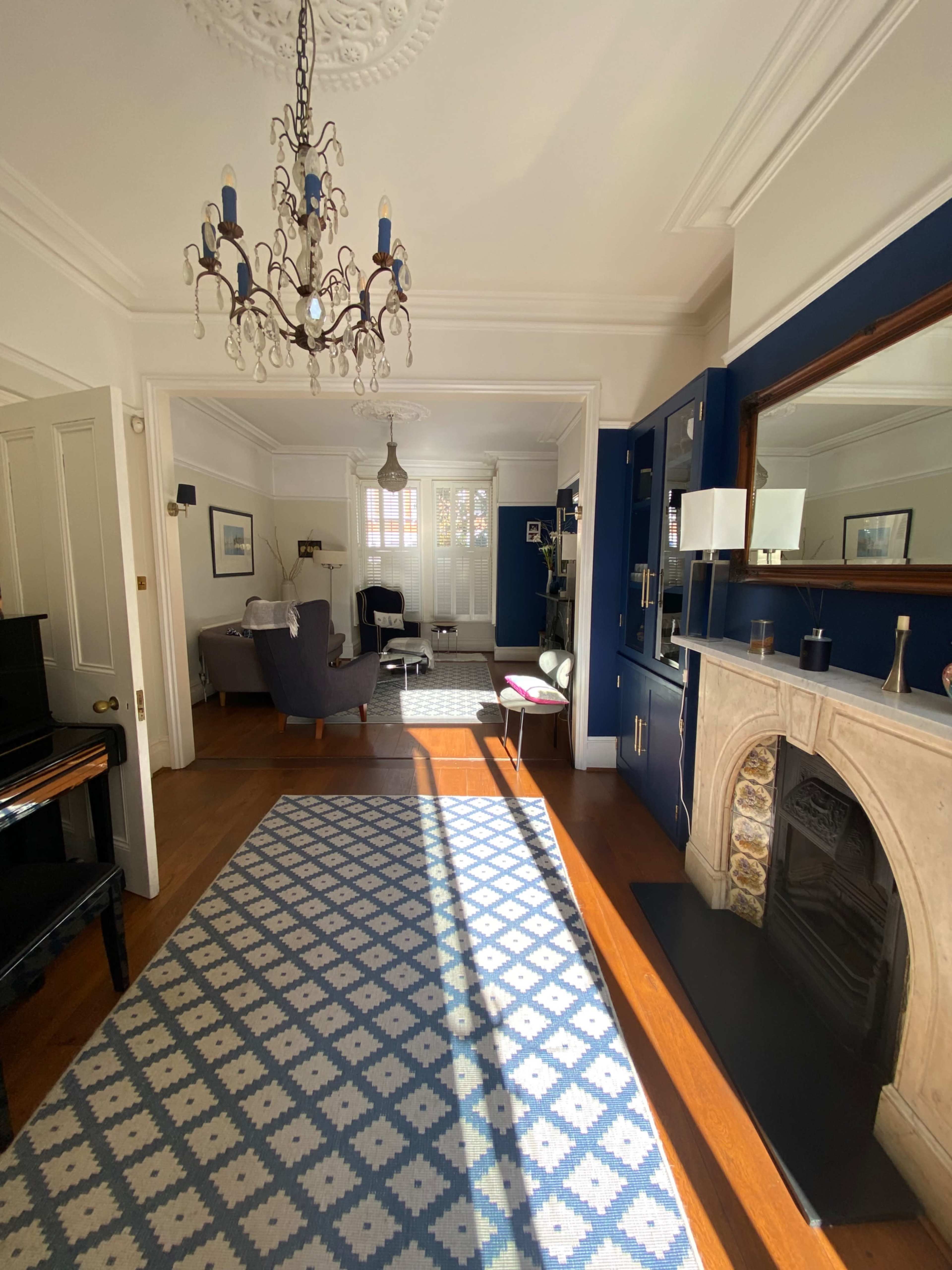 A well-lit living room features a patterned rug, a chandelier, a fireplace, and a spacious seating area with large windows.
