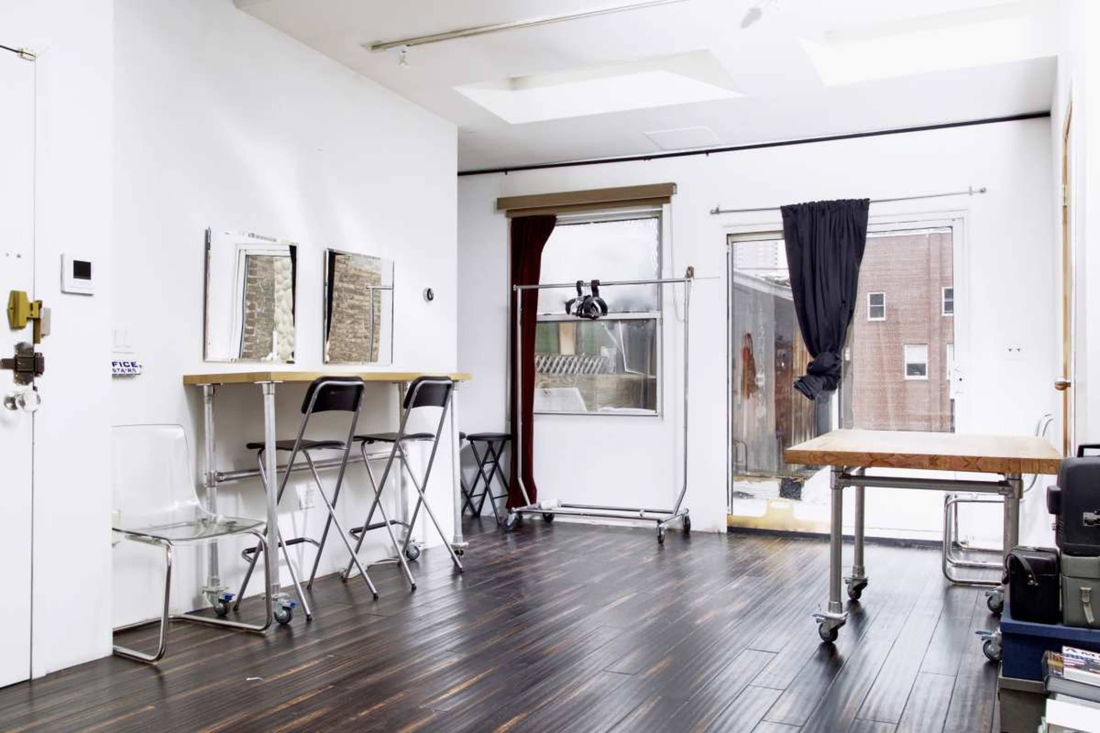 The image shows a bright, minimalistic room with large windows, two mirrors on a counter, a clothing rack, and a wooden table.