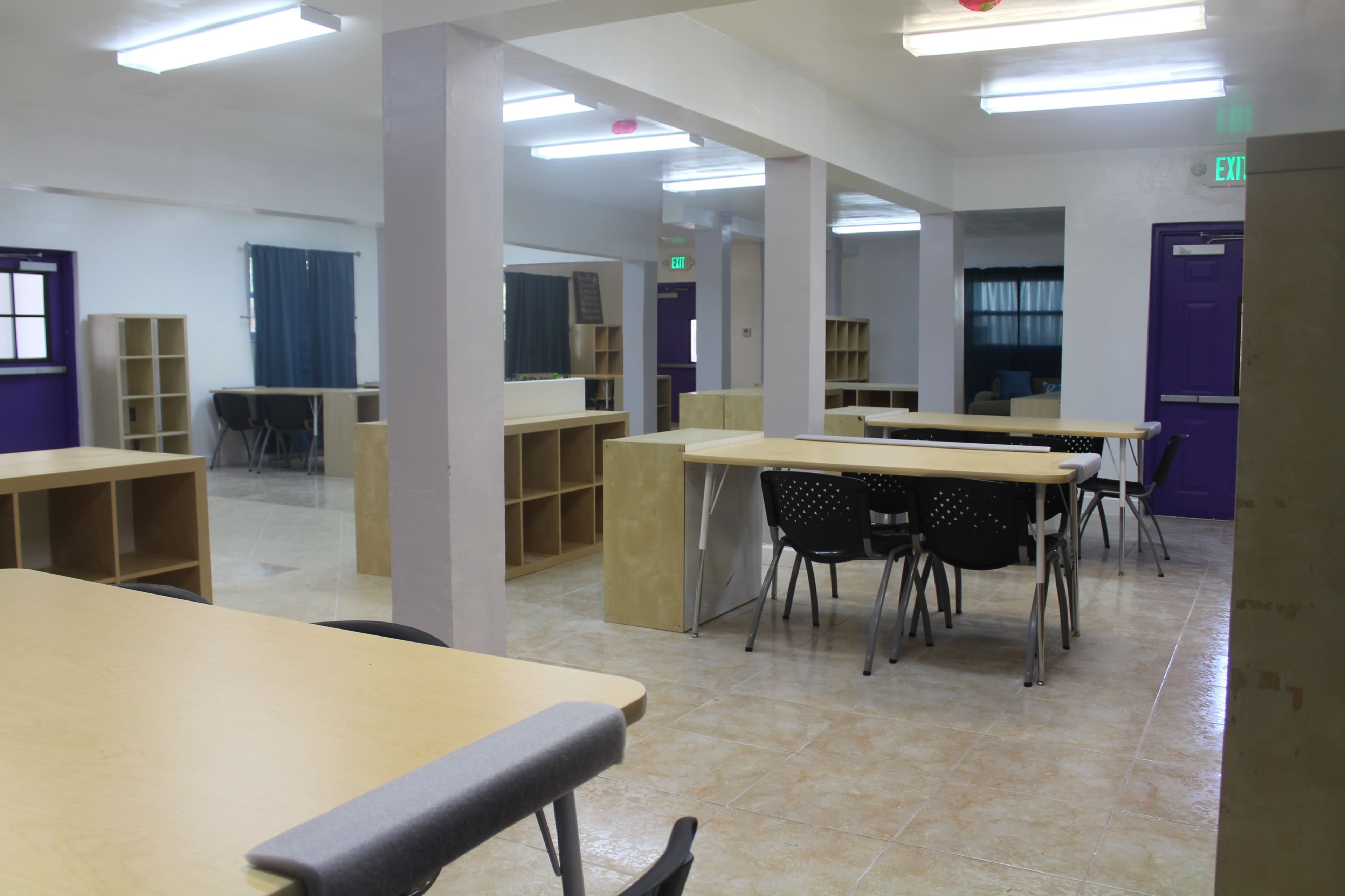 The image shows a brightly lit room with multiple wooden tables and black chairs, along with shelving units lining the walls.
