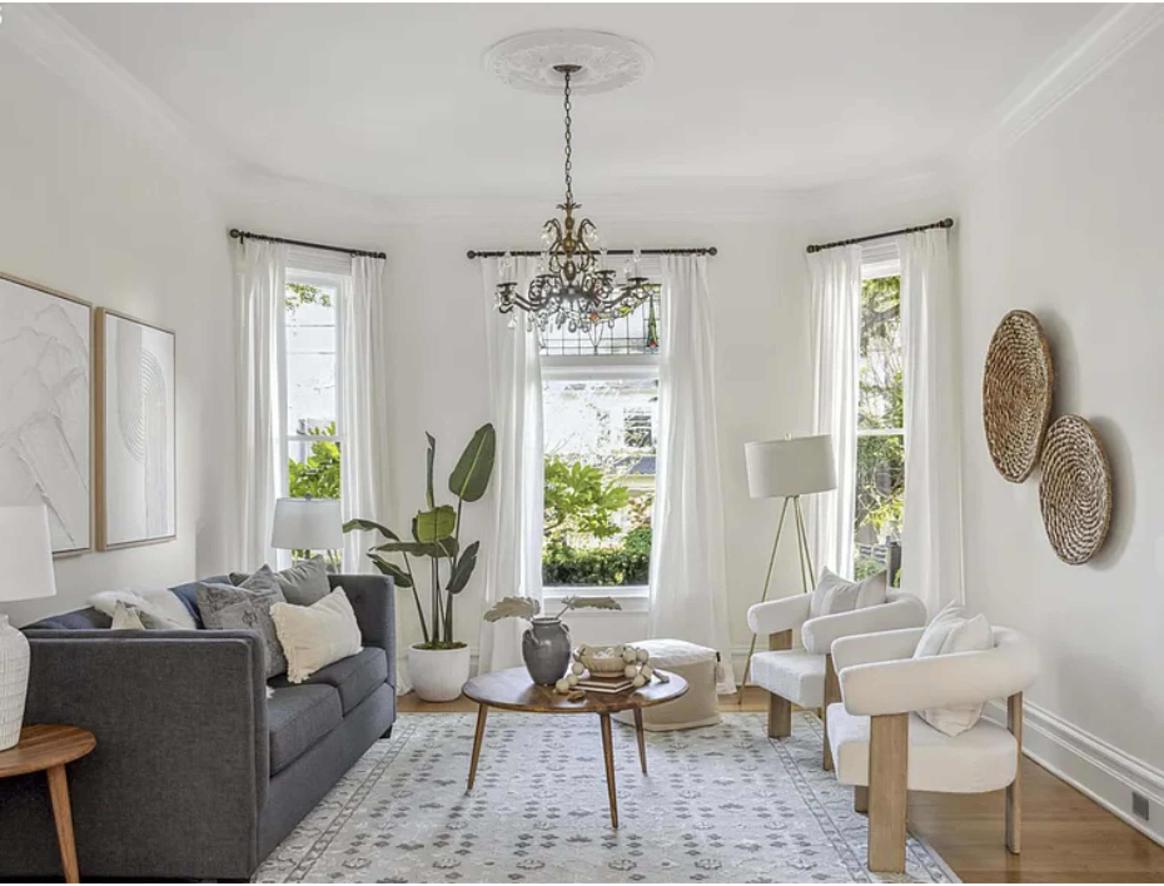 The living room features a gray sofa, a round coffee table, two white armchairs, and large windows with sheer curtains that let in natural light.