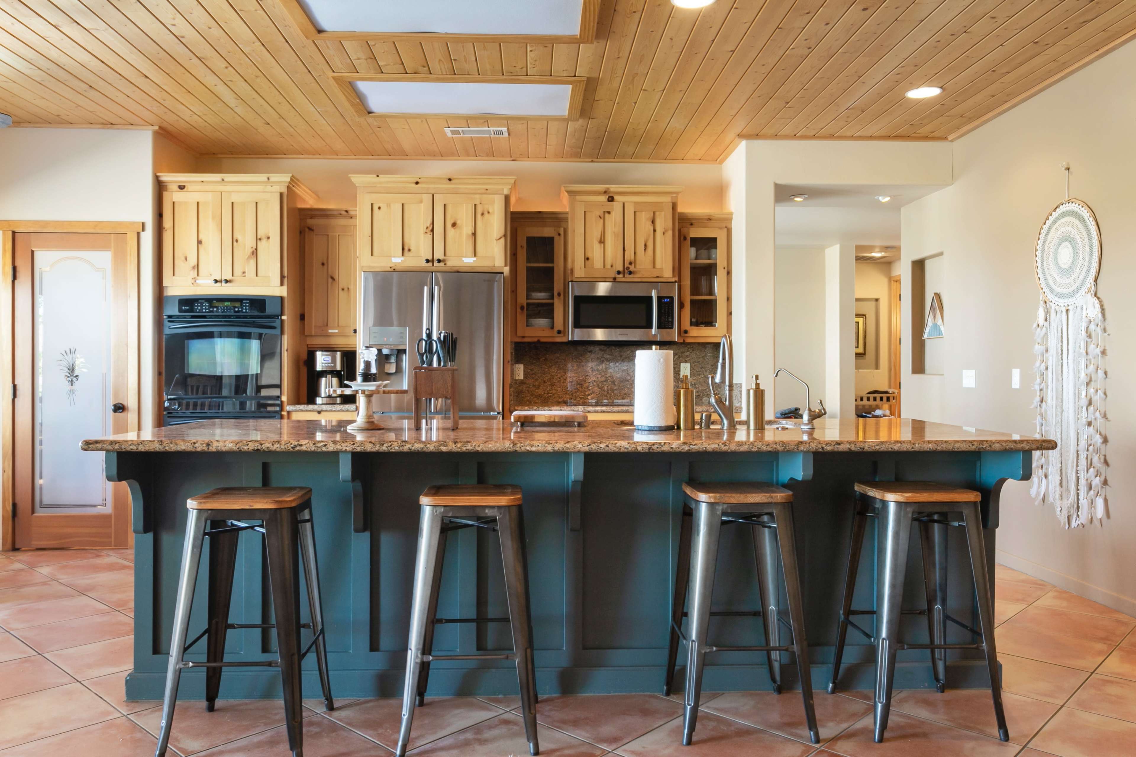 The image shows a modern kitchen with wooden cabinets, granite countertops, and four metal stools arranged at a central island.