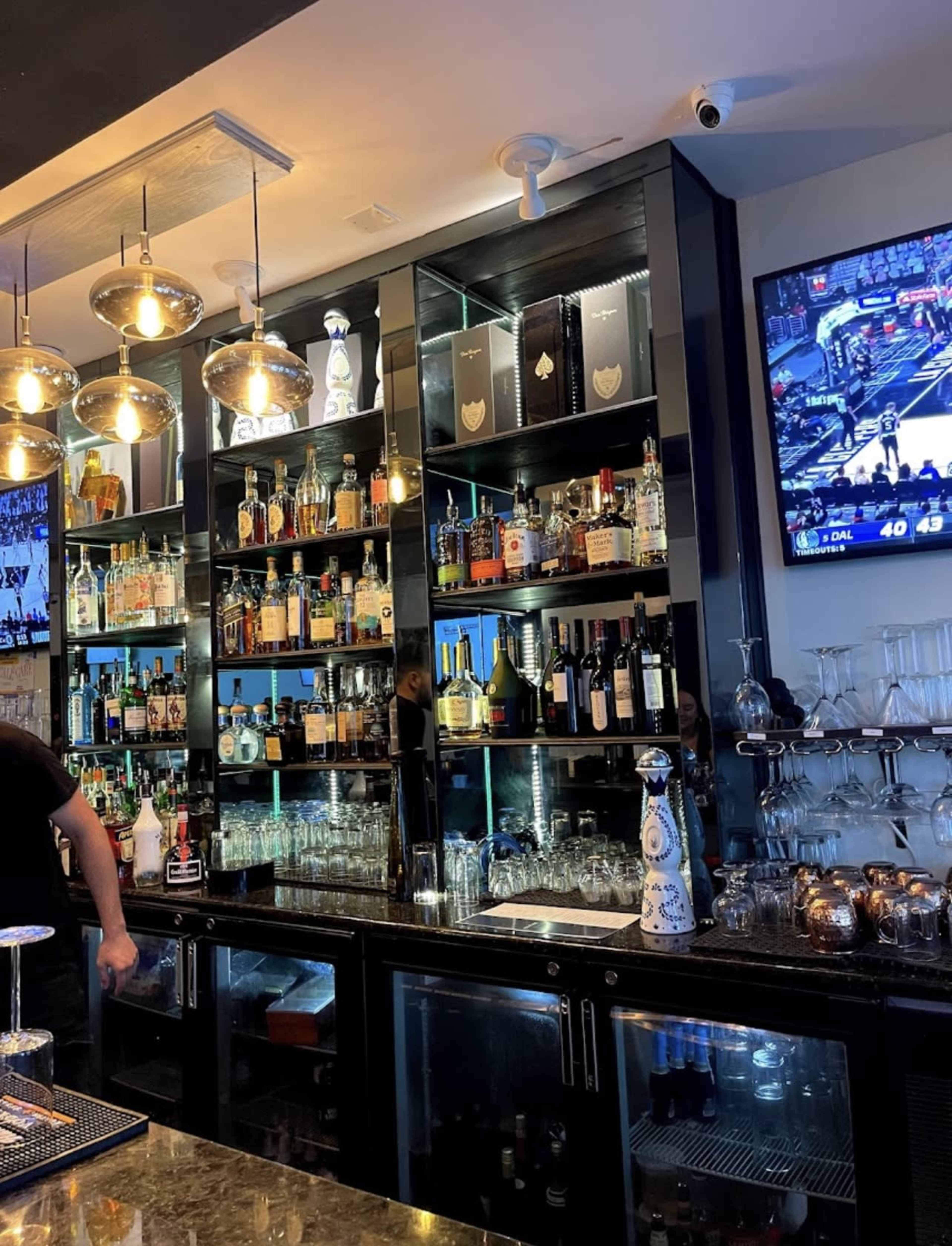 A bar counter displays an array of bottles and glassware, with a television screen showing a sports game in the background.