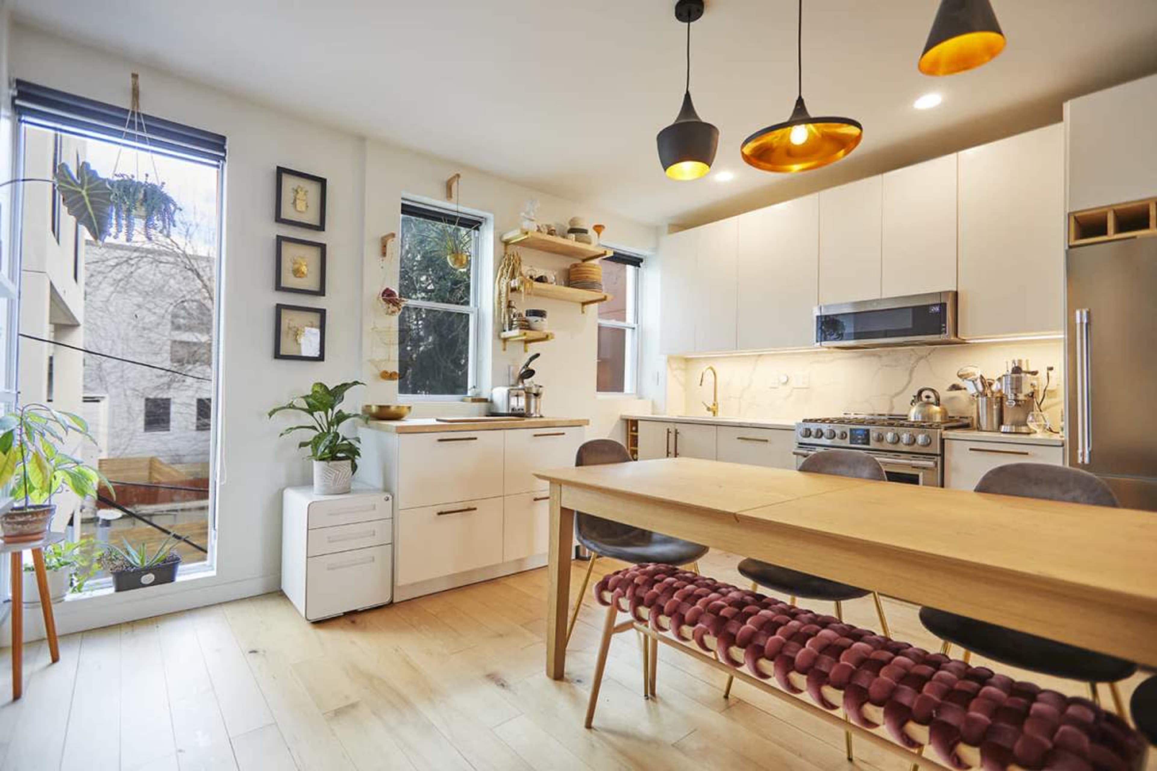 A modern kitchen features a wooden dining table, sleek cabinetry, and large windows allowing natural light.