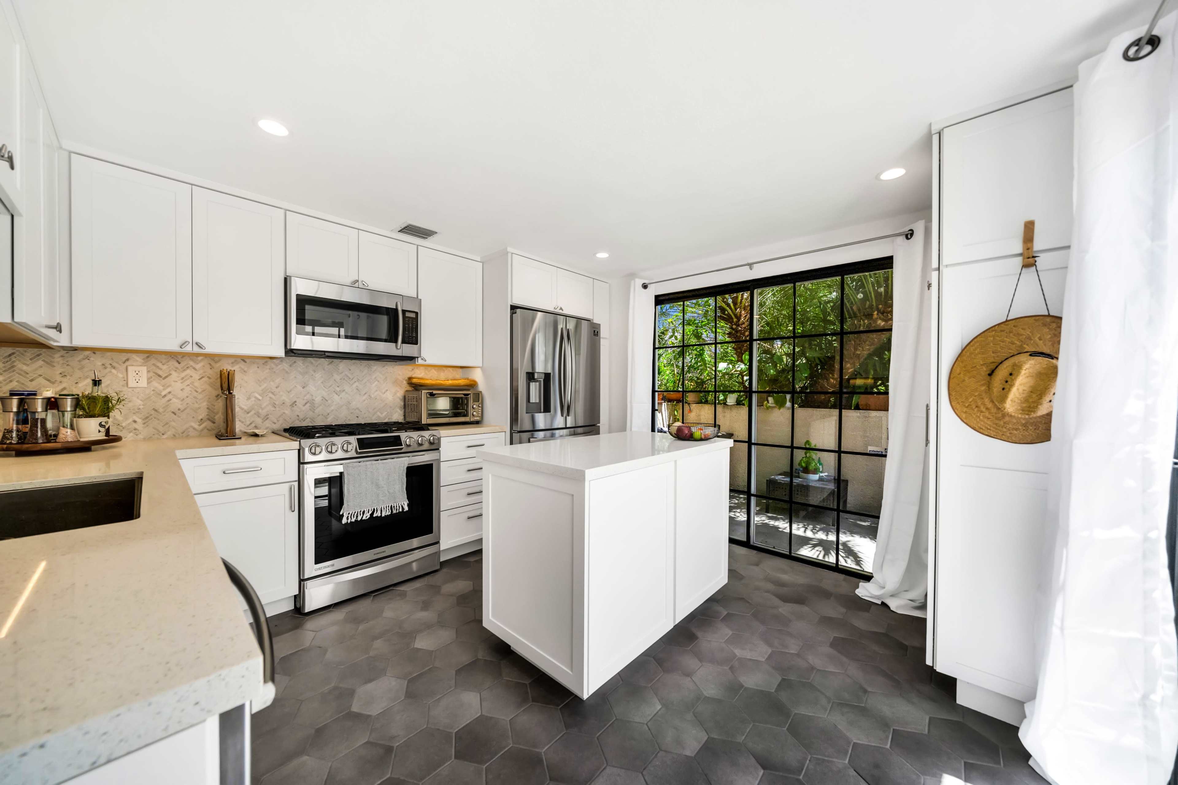 A modern kitchen features white cabinetry, a stainless steel oven, and a central island with a hexagonal tile floor and a window overlooking a garden.