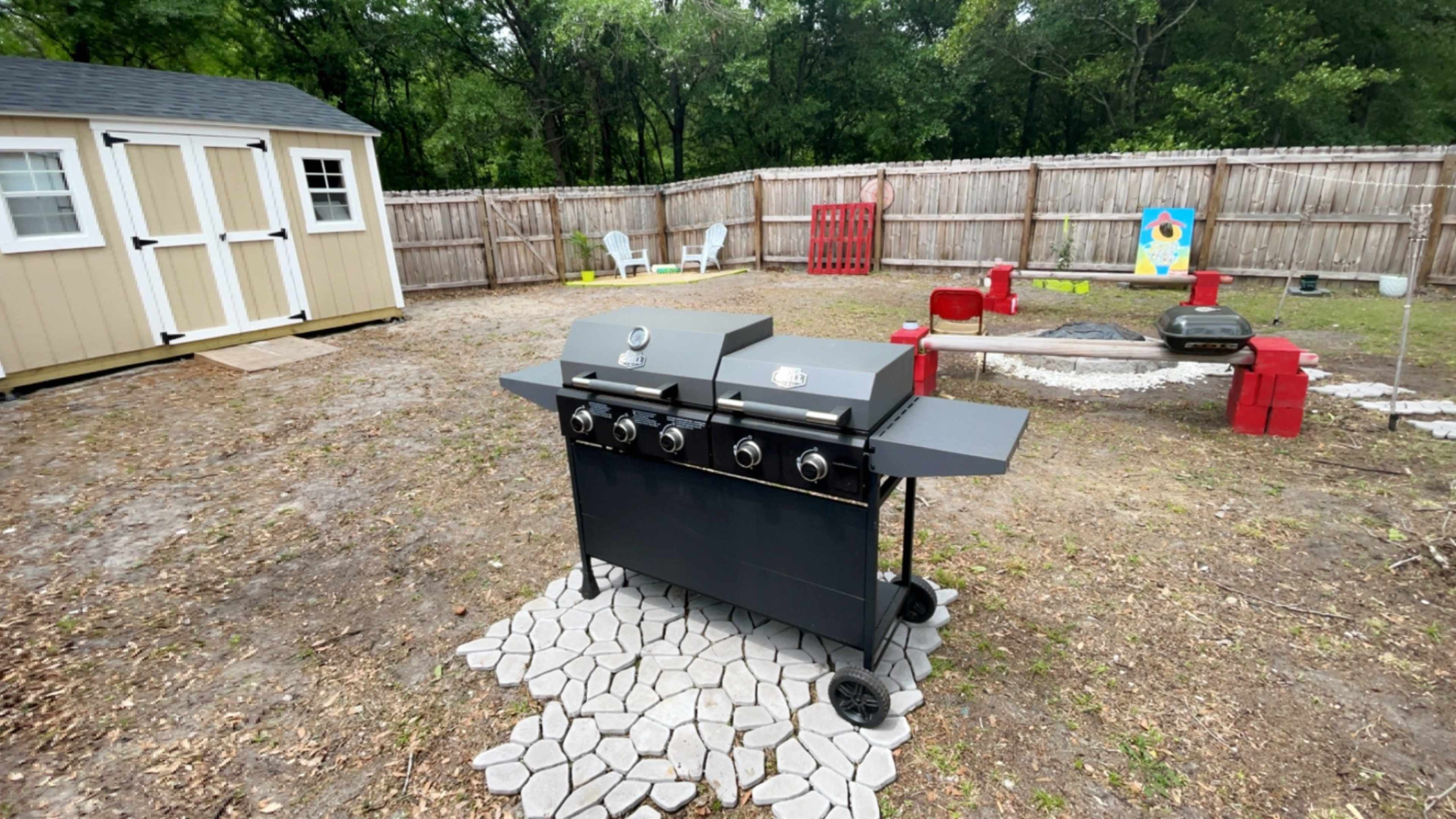 A black gas grill sits on a stone patio in an outdoor backyard, surrounded by a wooden fence and various lawn furniture.