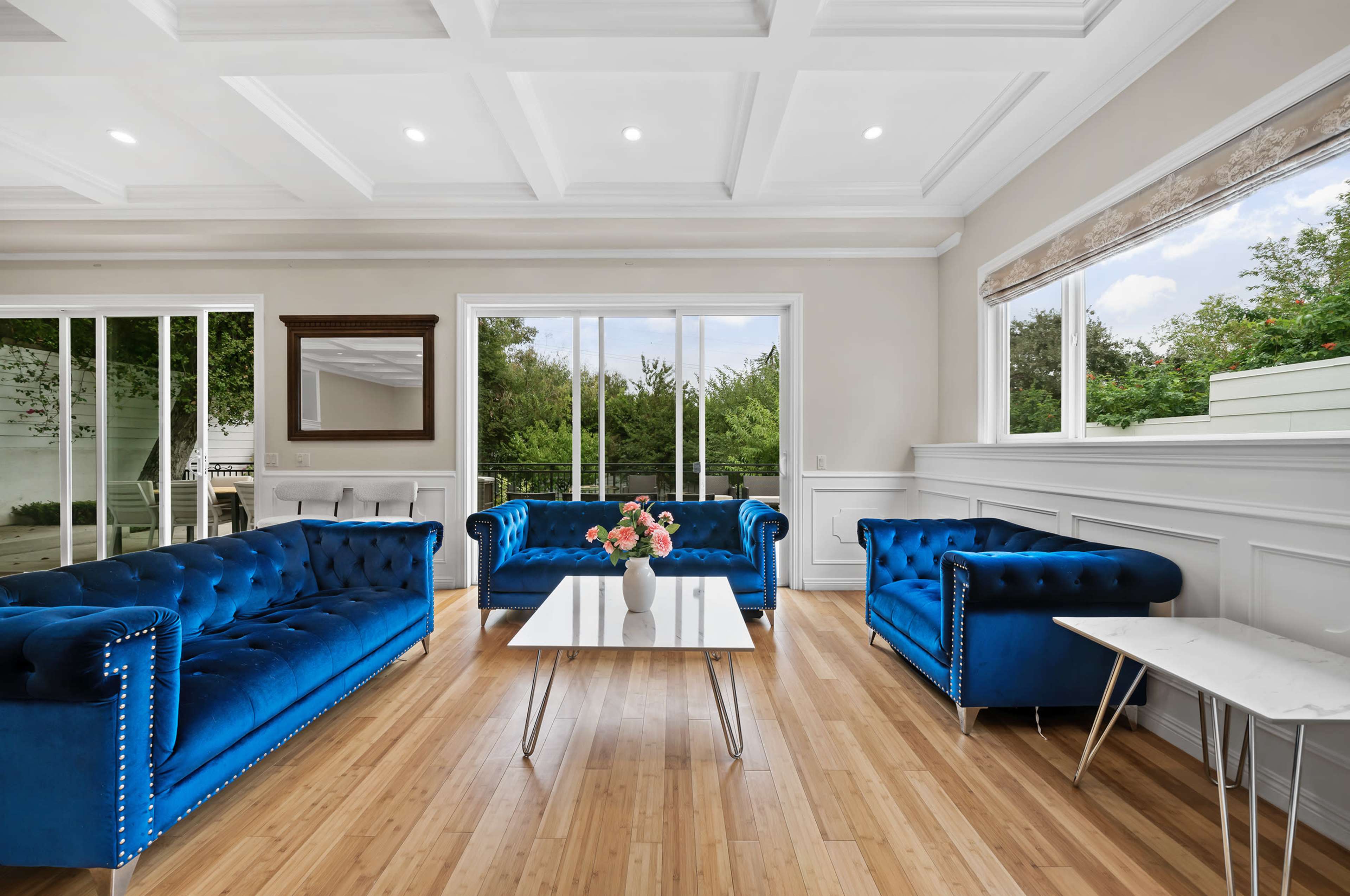 The living room features two blue velvet sofas facing a glass coffee table, with large windows overlooking greenery.