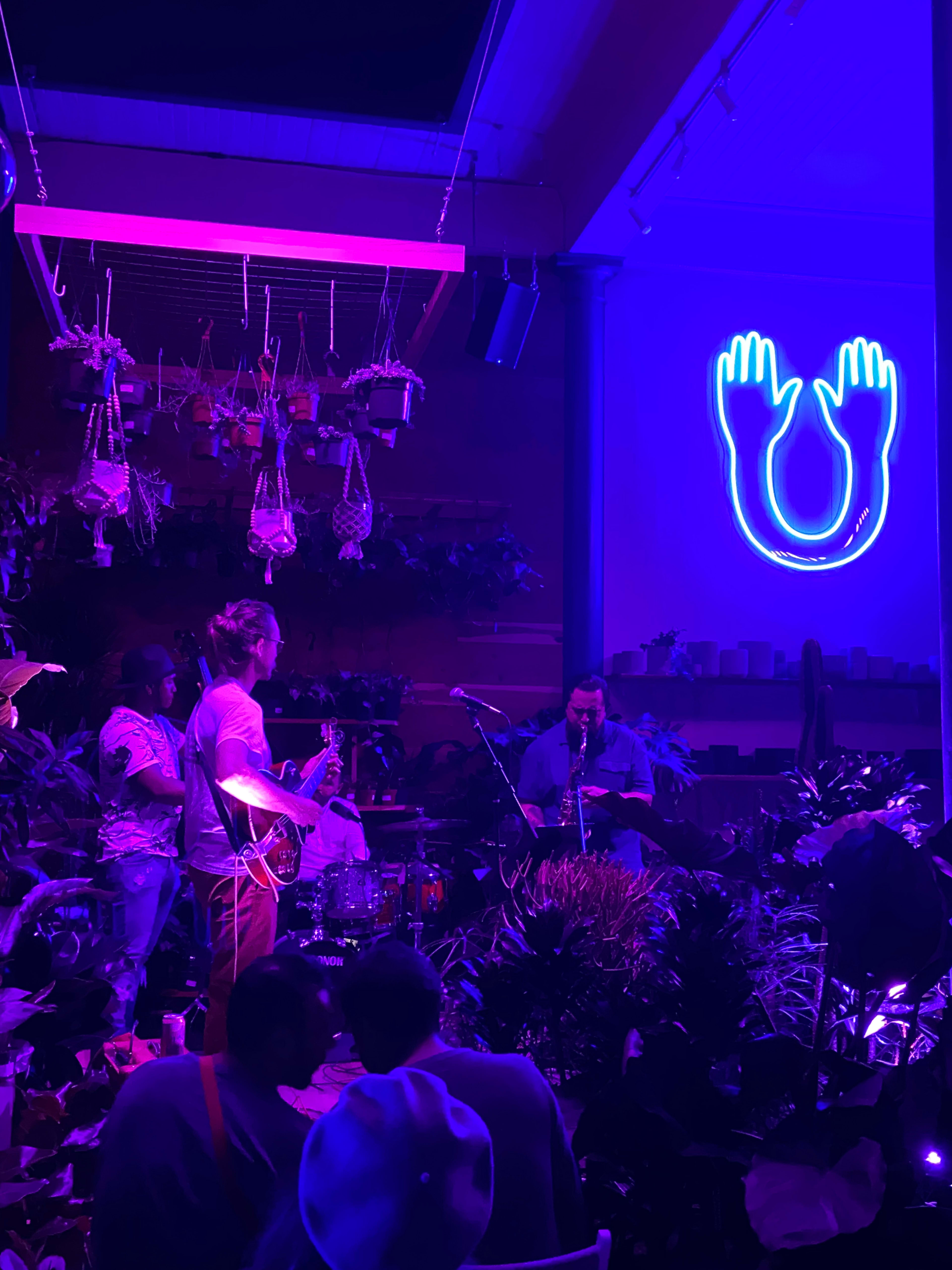A band performs on stage in a dimly lit venue filled with plants, while a neon blue sign of hands and a horseshoe illuminates the background.