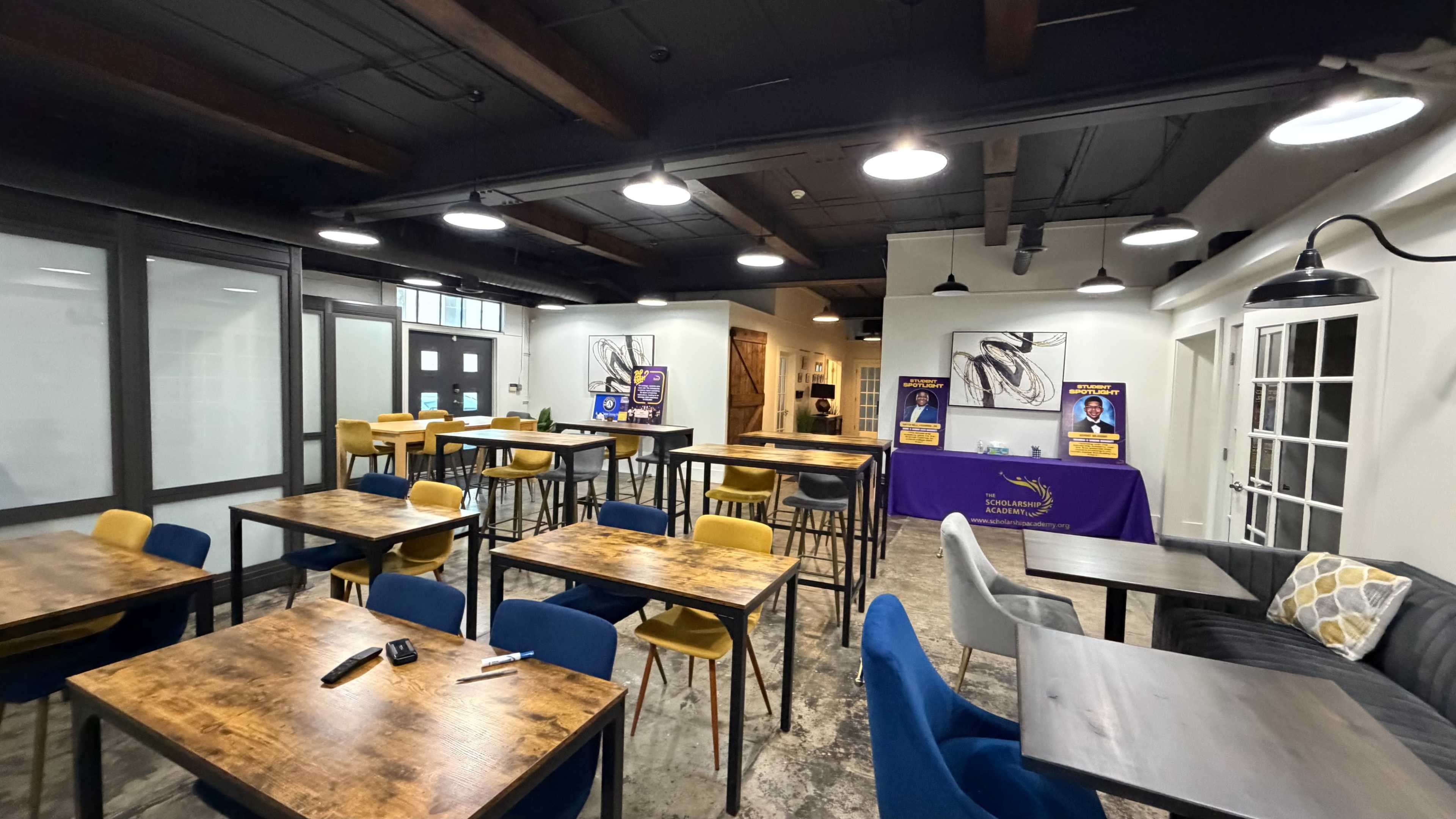 The image shows a modern interior of a café or meeting space featuring wooden tables and colorful chairs, with promotional materials on a table at the back.