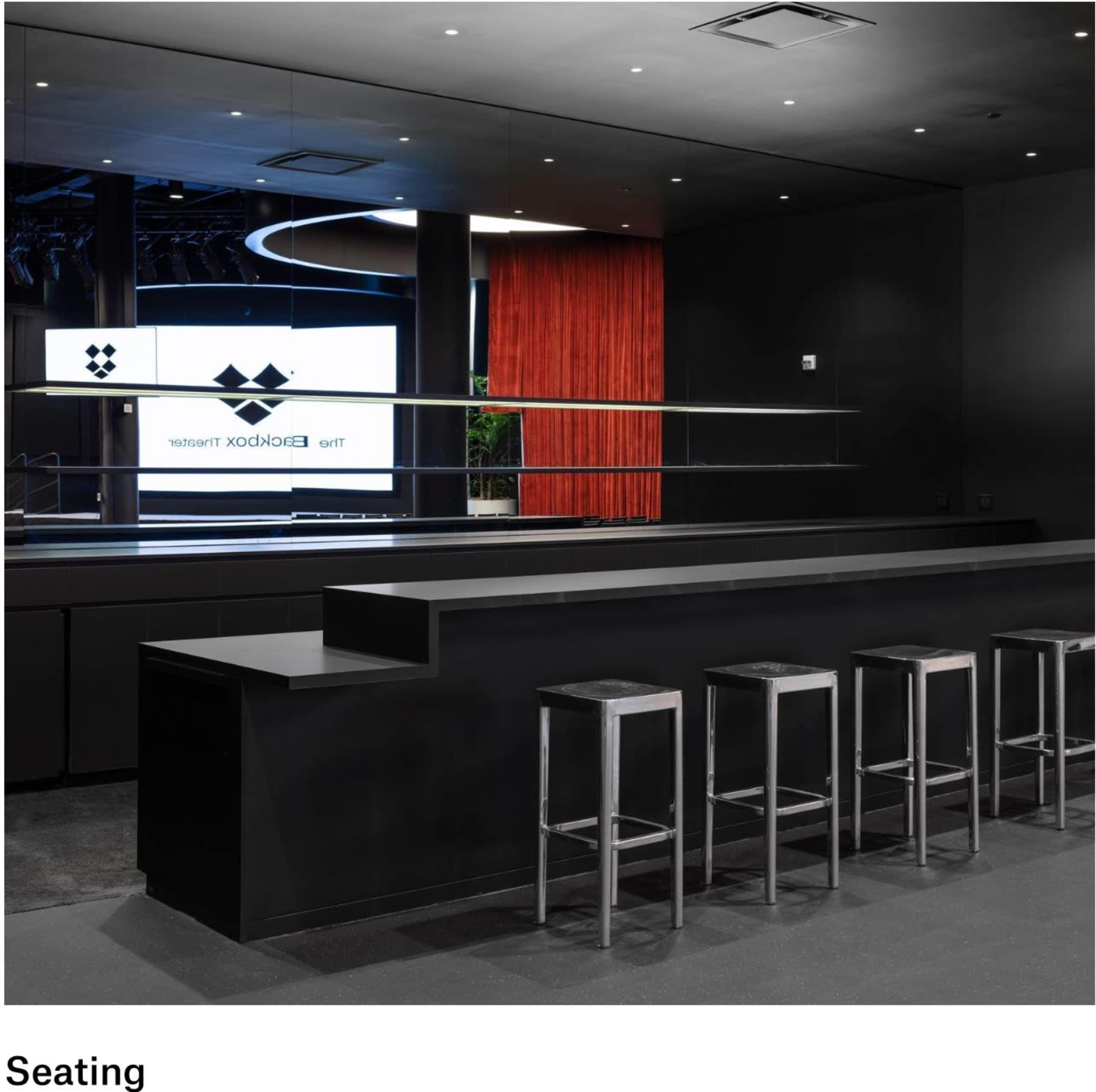 A modern bar area features sleek black furniture, including high stools aligned at a counter, with a backdrop of a large screen and red drapery.