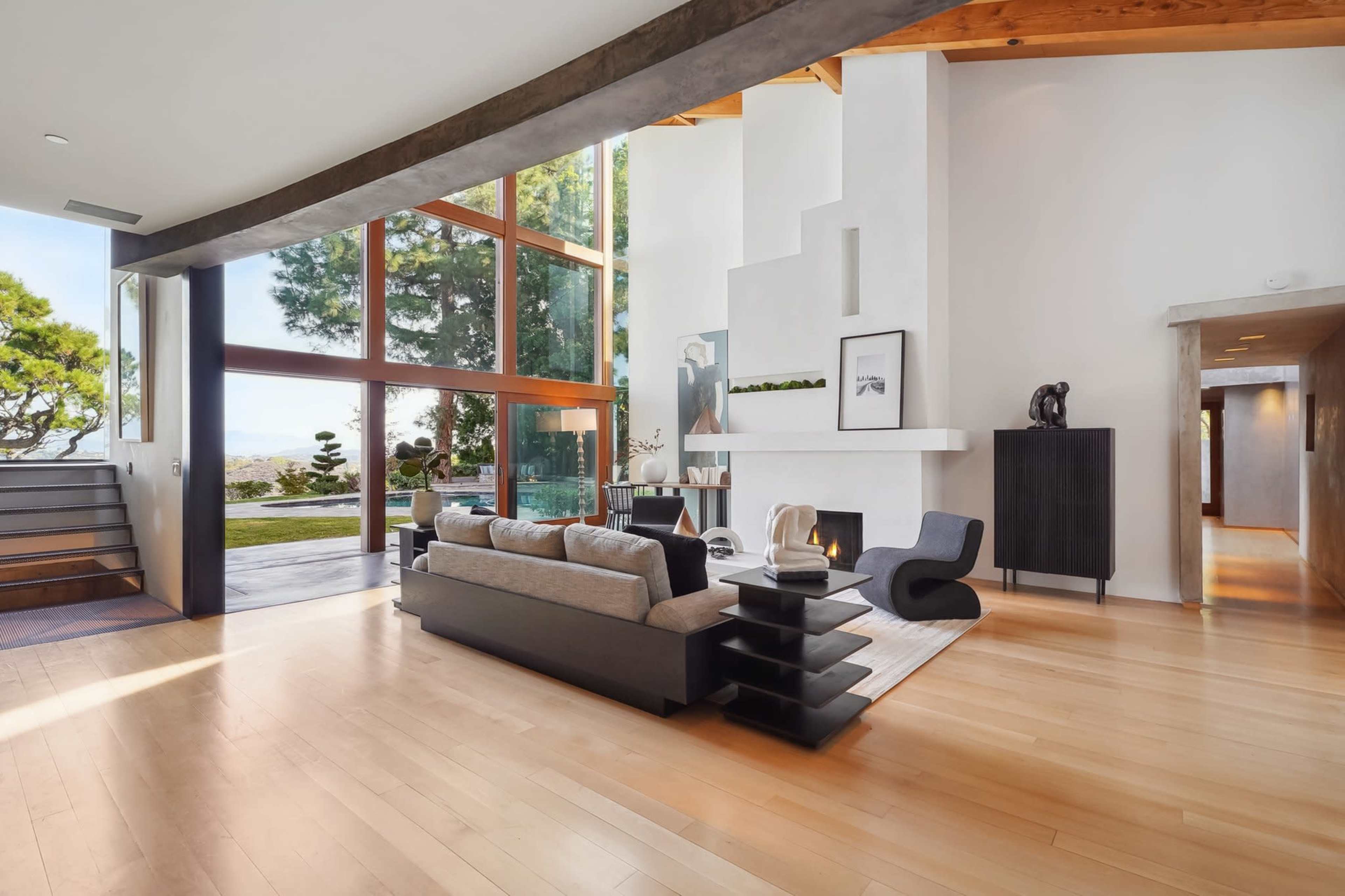 The image shows a modern living room with large windows, a fireplace, a sleek sofa, and an open floor plan leading to an outdoor view.
