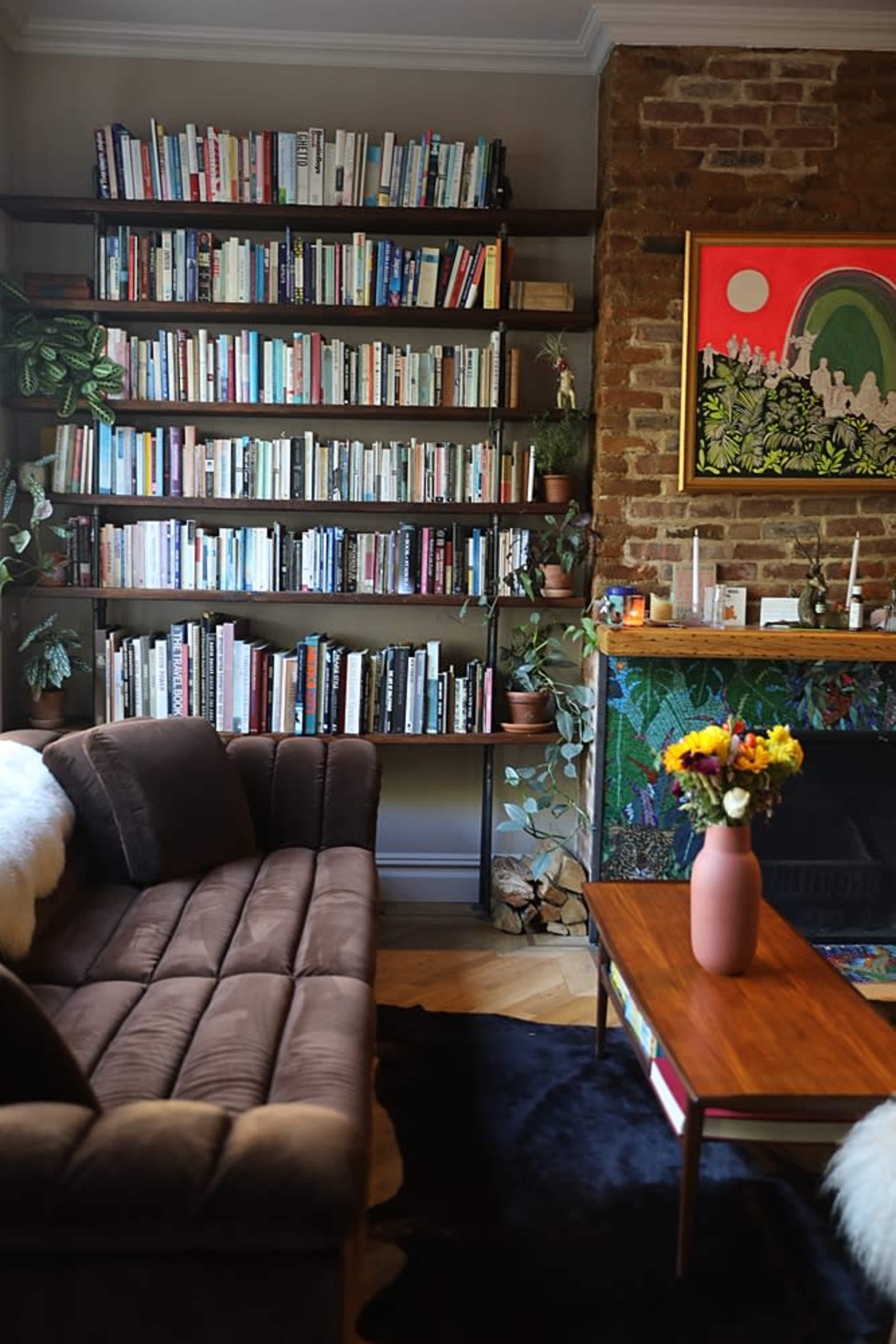 A cozy living room features a brown sofa, a wooden coffee table with a vase of flowers, and a wall of bookshelves filled with various books and plants.