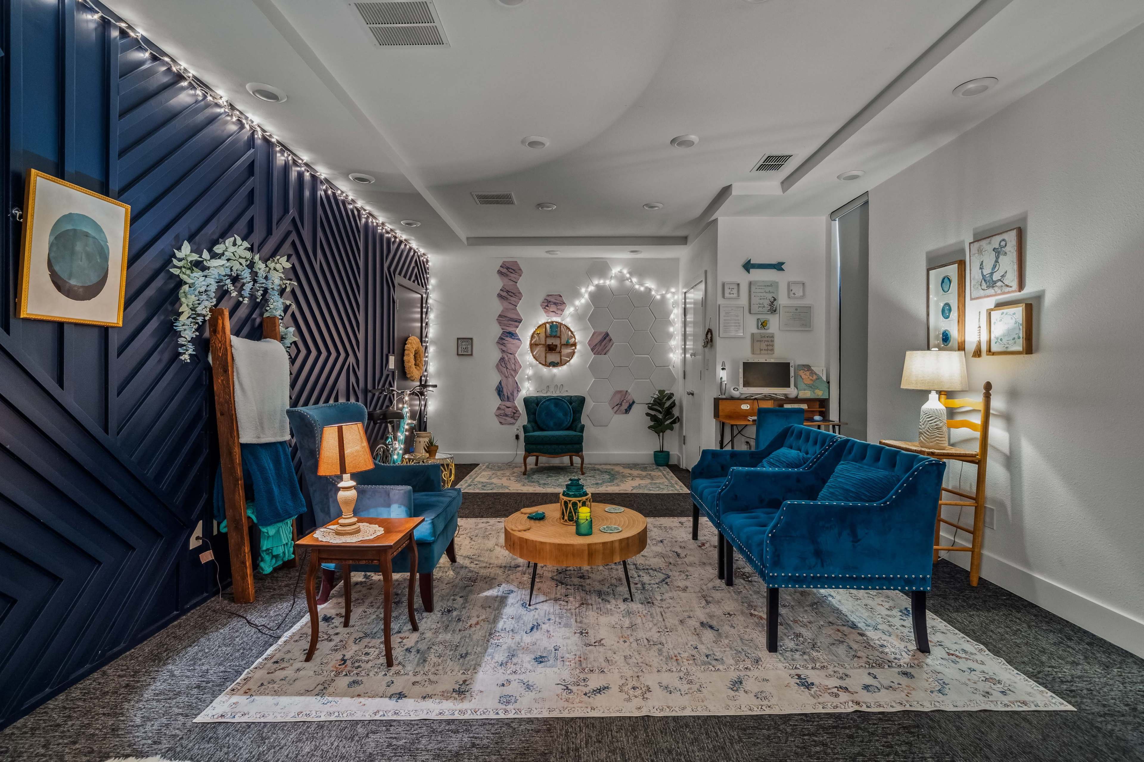 The image shows a cozy lounge area with blue upholstered chairs, a round wooden coffee table, and decorative wall accents including artwork and soft lighting.