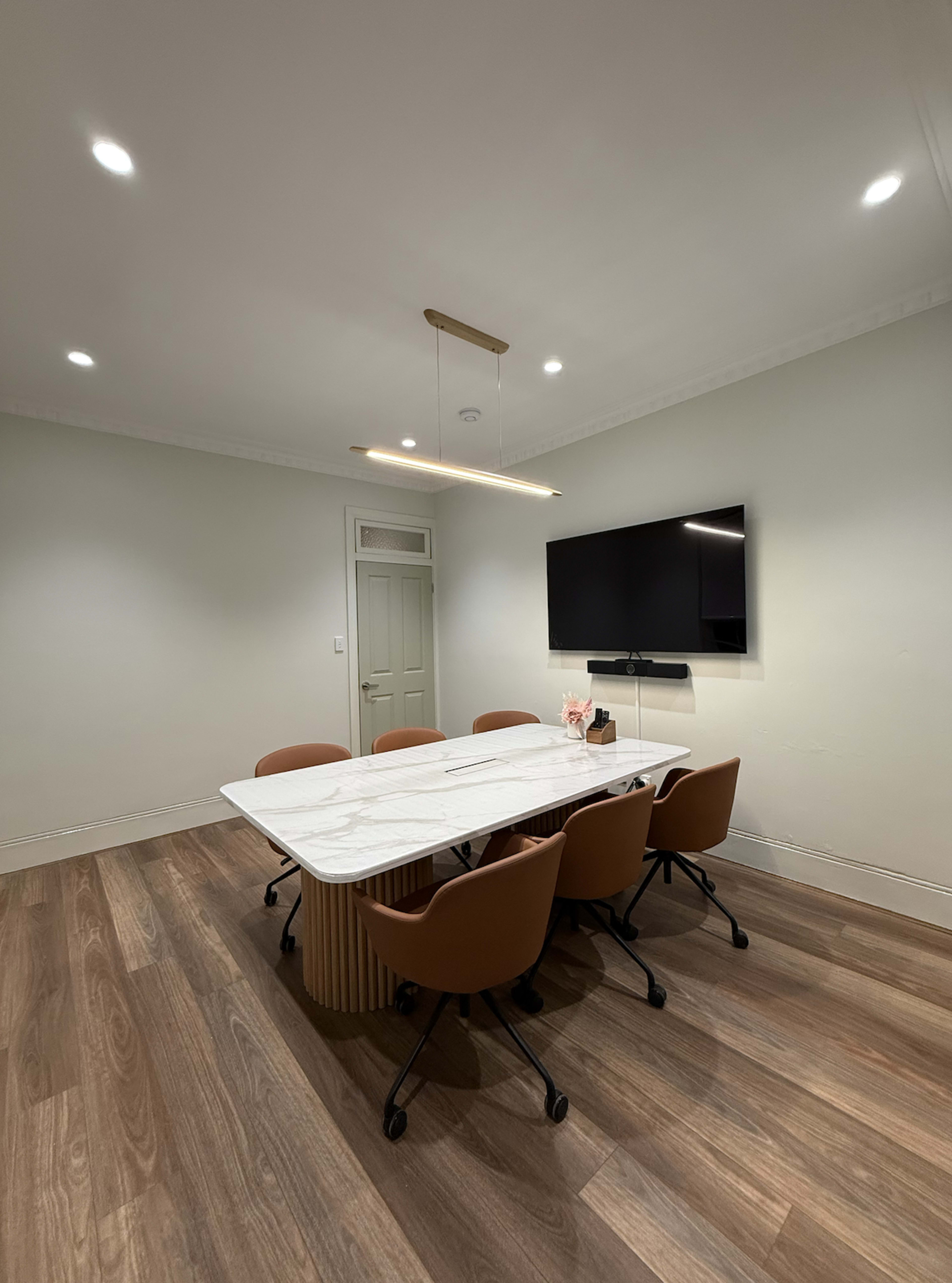 The image shows a modern conference room with a marble-topped table surrounded by four brown chairs, a wall-mounted television, and light-colored walls.