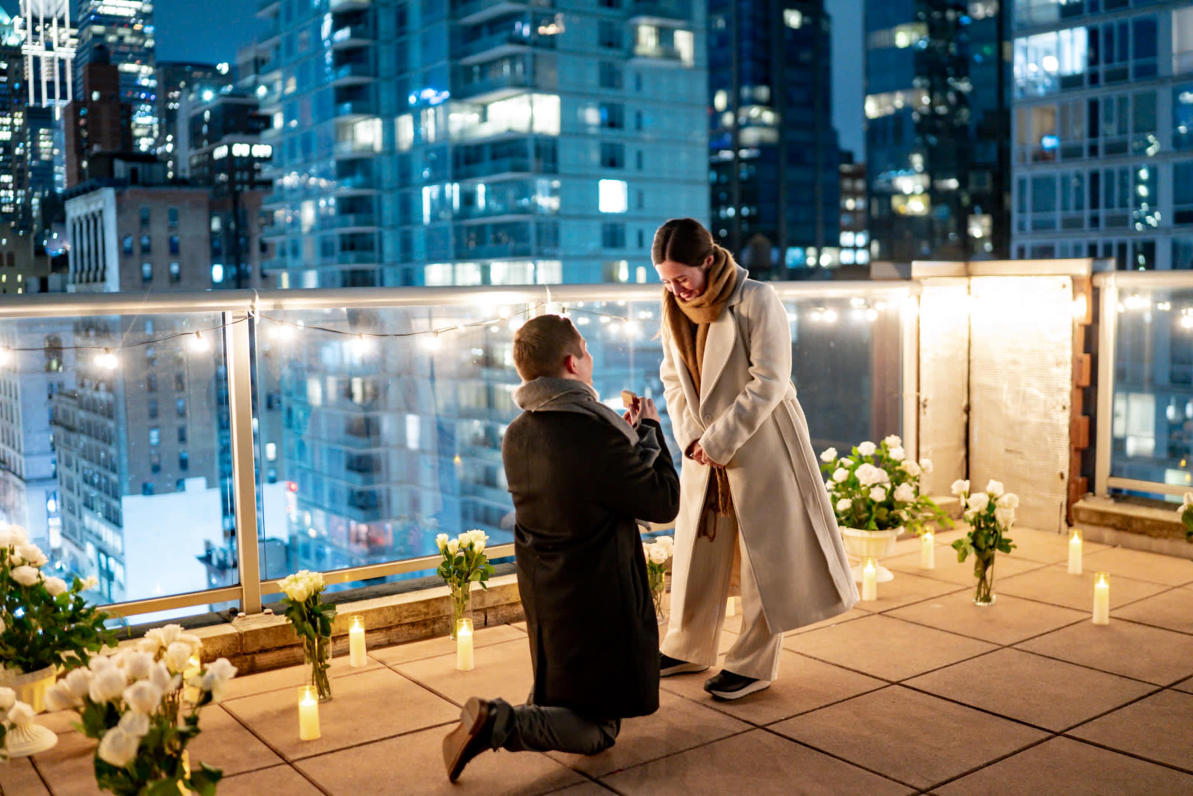 A man is proposing to a woman on a city rooftop, surrounded by white roses and candles, with a skyline in the background.