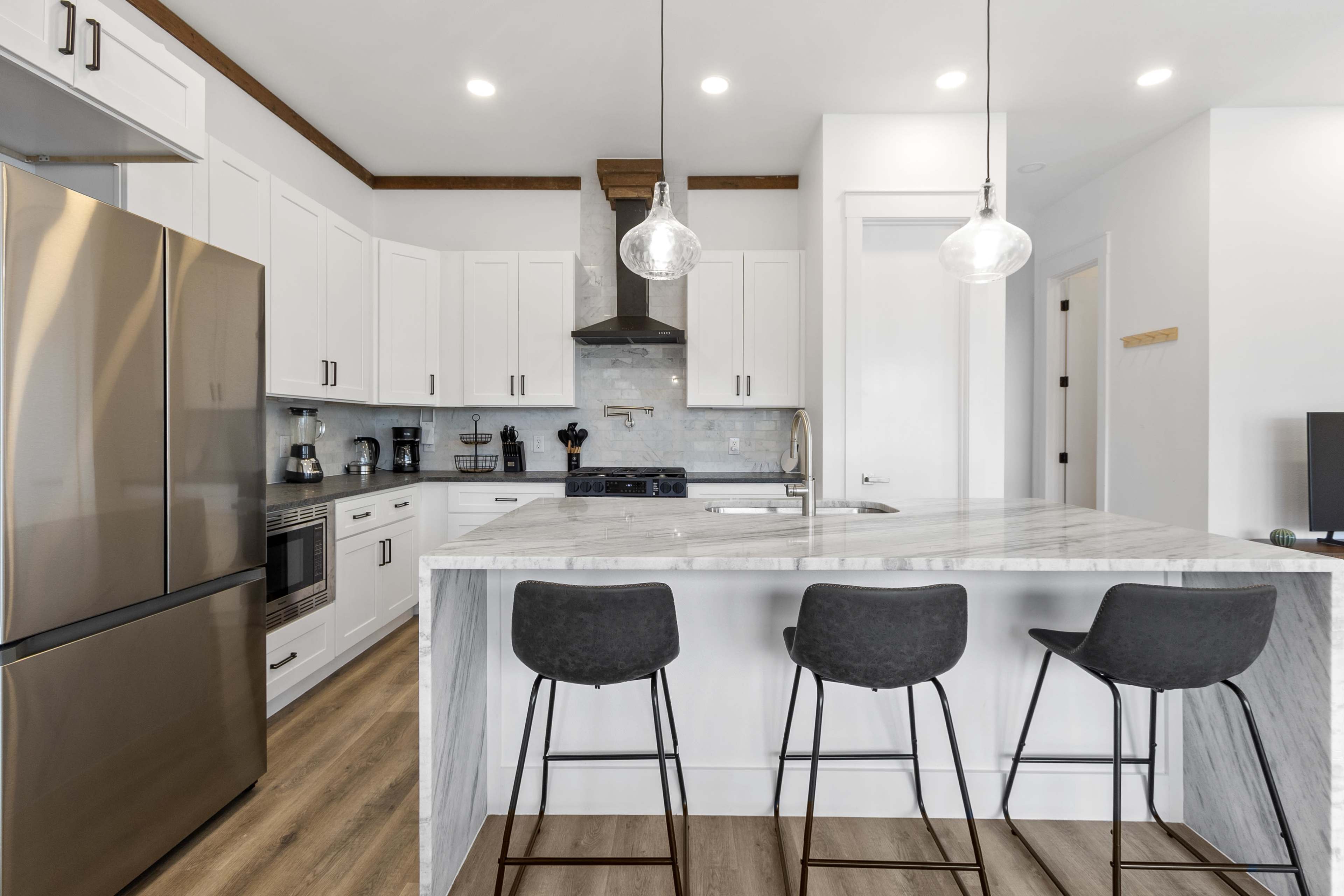 The image shows a modern kitchen with white cabinetry, stainless steel appliances, and a marble-topped island featuring three bar stools.