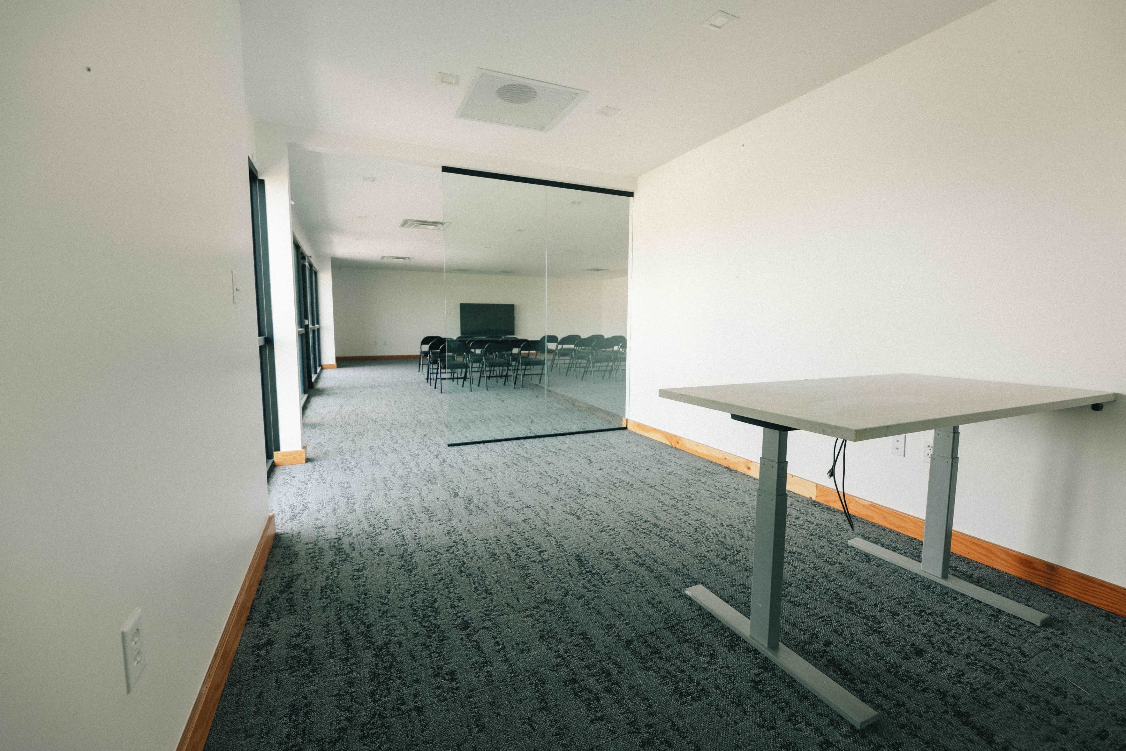 The image shows a modern conference room with a glass wall, empty chairs arranged in a section, and a moveable table in the foreground.