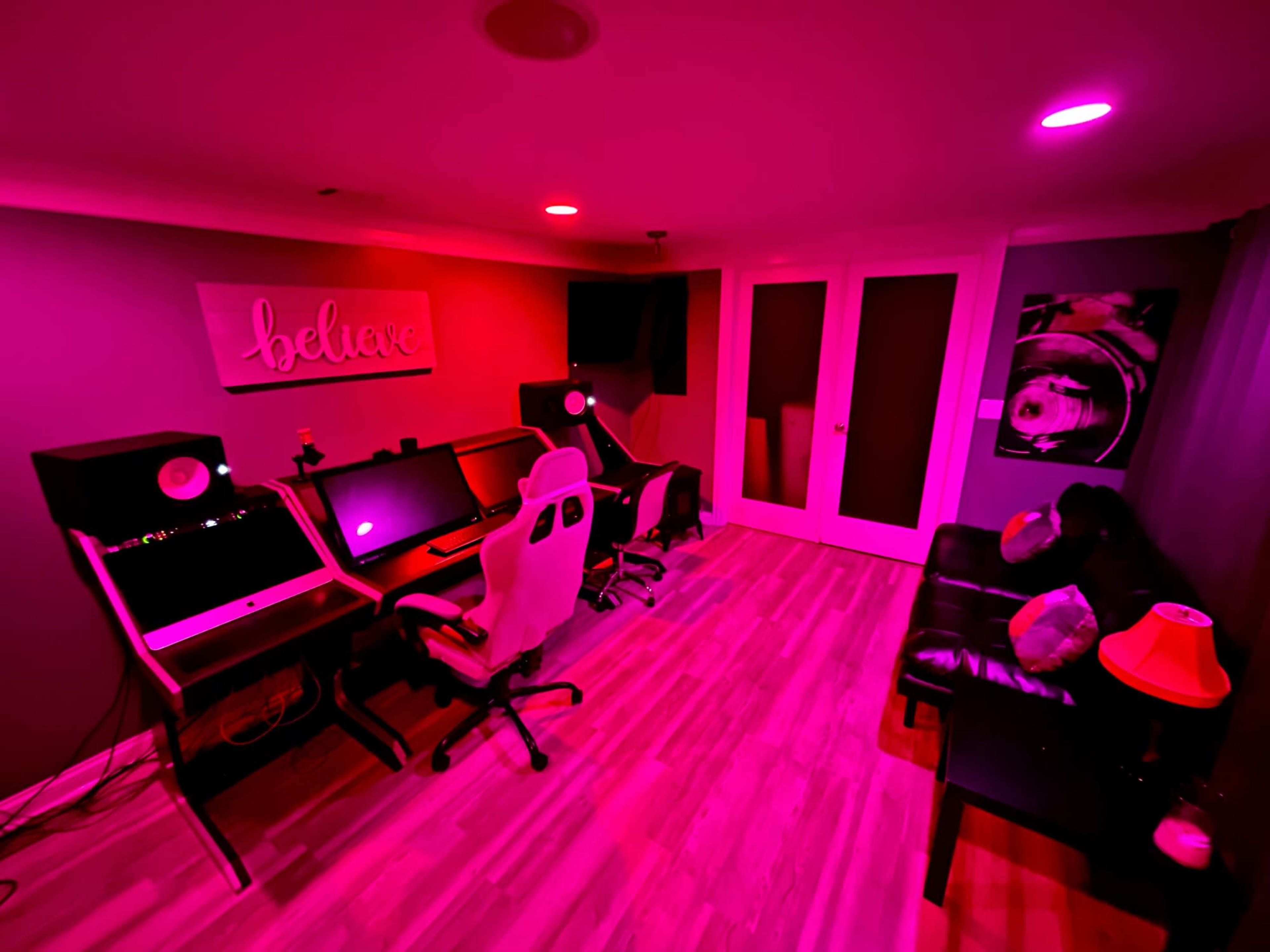 A modern home studio setup features two computer desks with monitors, audio speakers, and gaming chairs in a room illuminated by pink lighting.