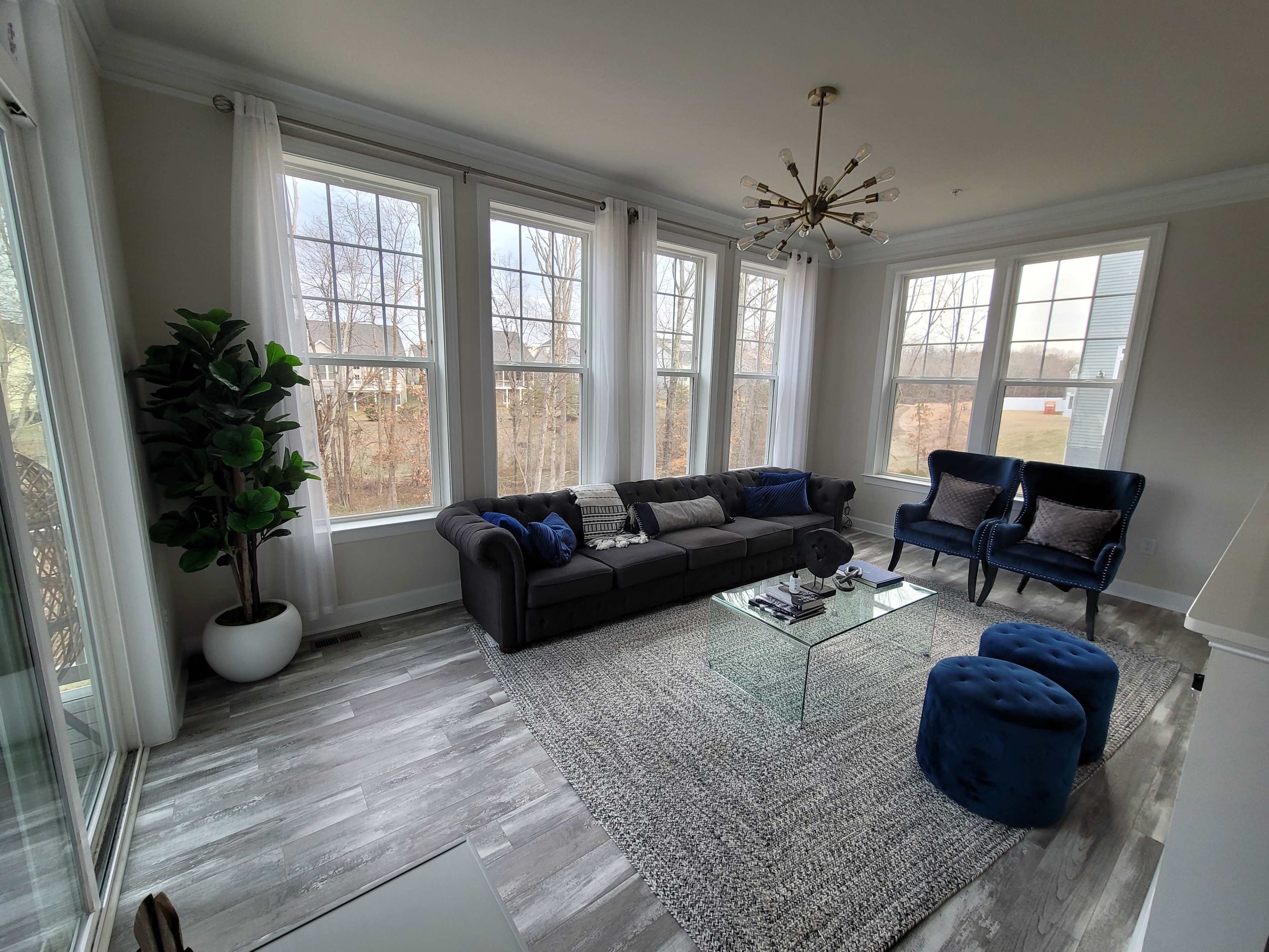 A modern living room features large windows, a gray couch with blue pillows, a glass coffee table, and a blue ottoman, set against a backdrop of trees outside.