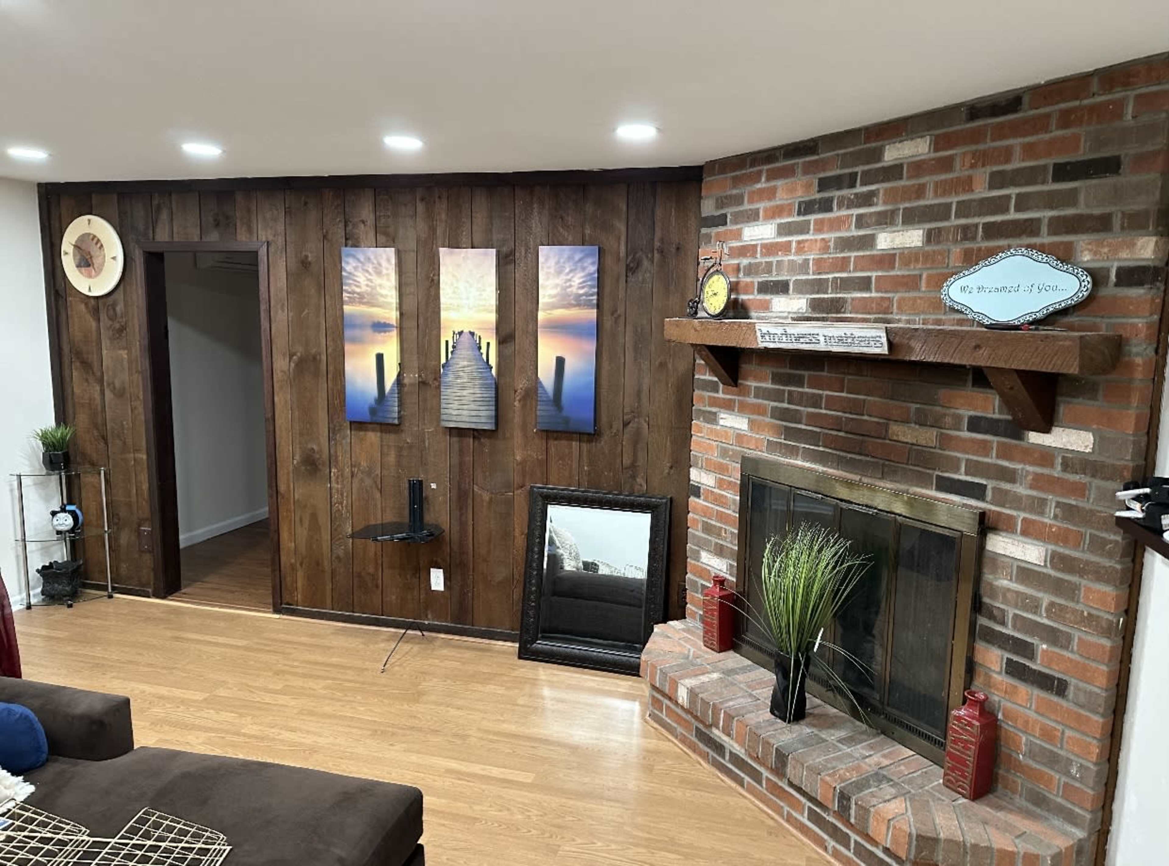 A living room with a wood-paneled wall featuring three landscape panels, a brick fireplace, and a shelf adorned with decorative items.