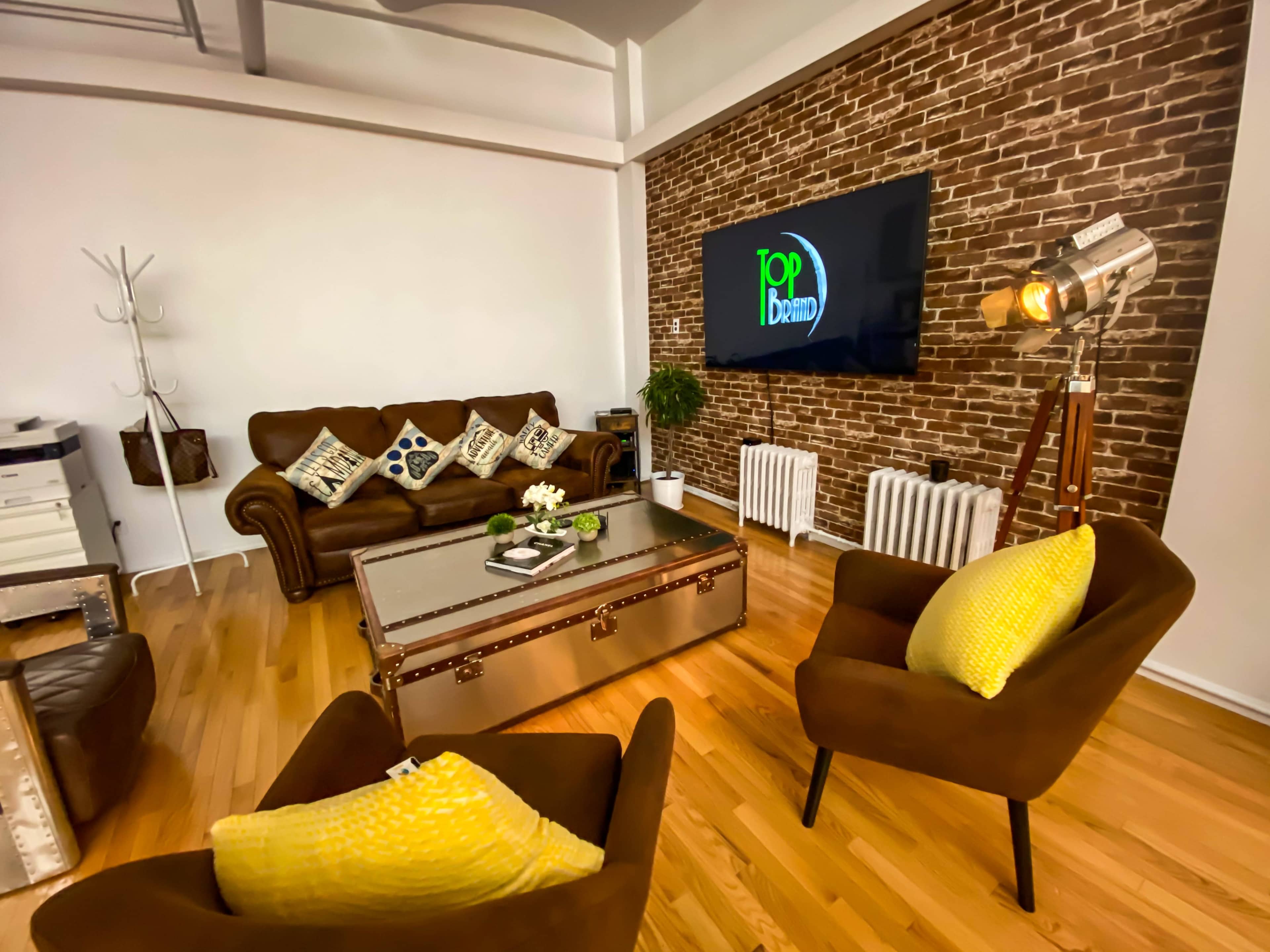 A modern living room featuring a brown leather sofa with patterned cushions, a vintage-style coffee table, two yellow accent chairs, and a large wall-mounted screen against a brick backdrop.