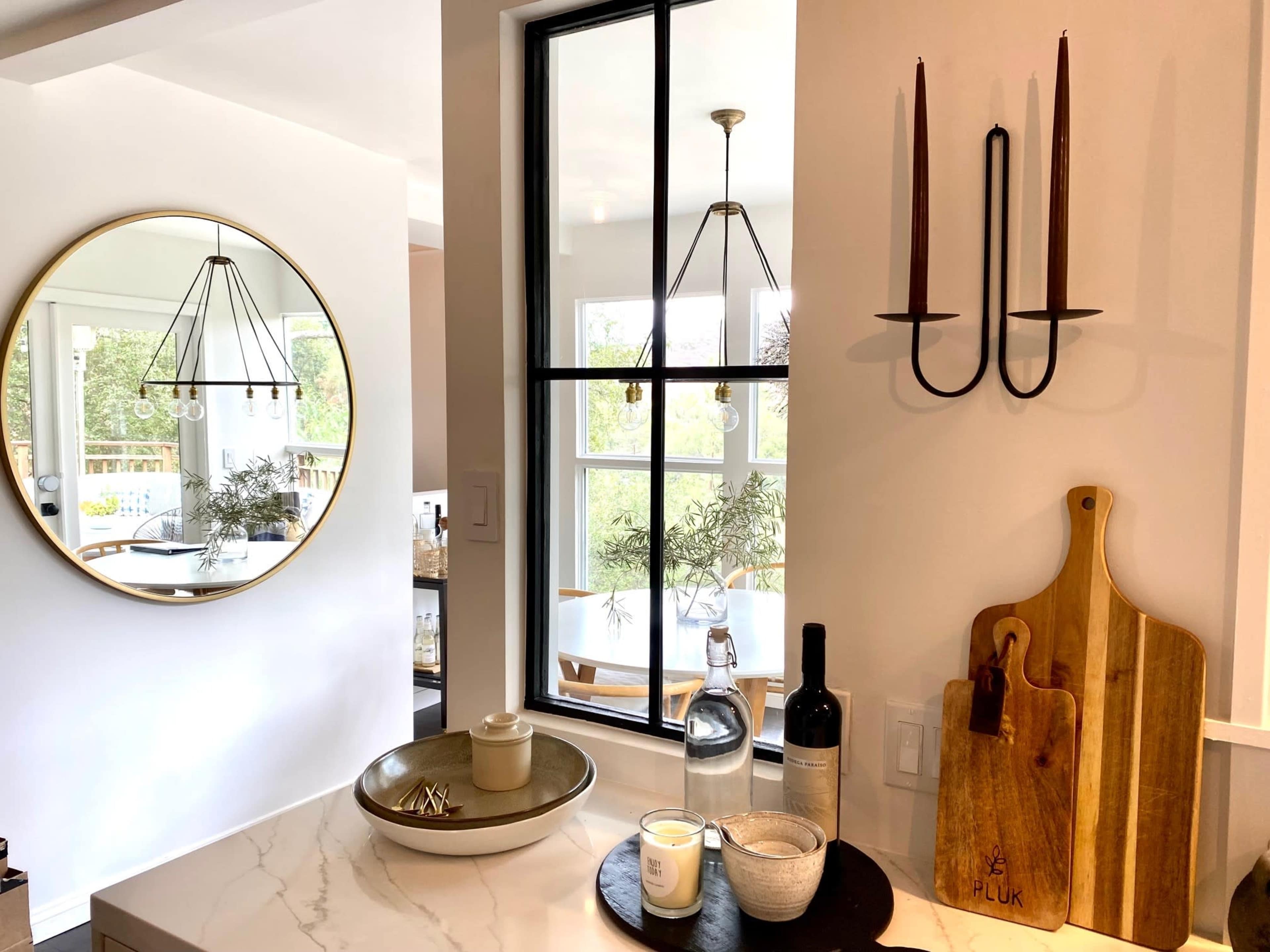 A kitchen scene features a round mirror, a candle on a plate, a bottle of wine, and a wooden cutting board next to a window with two candles mounted on the wall.