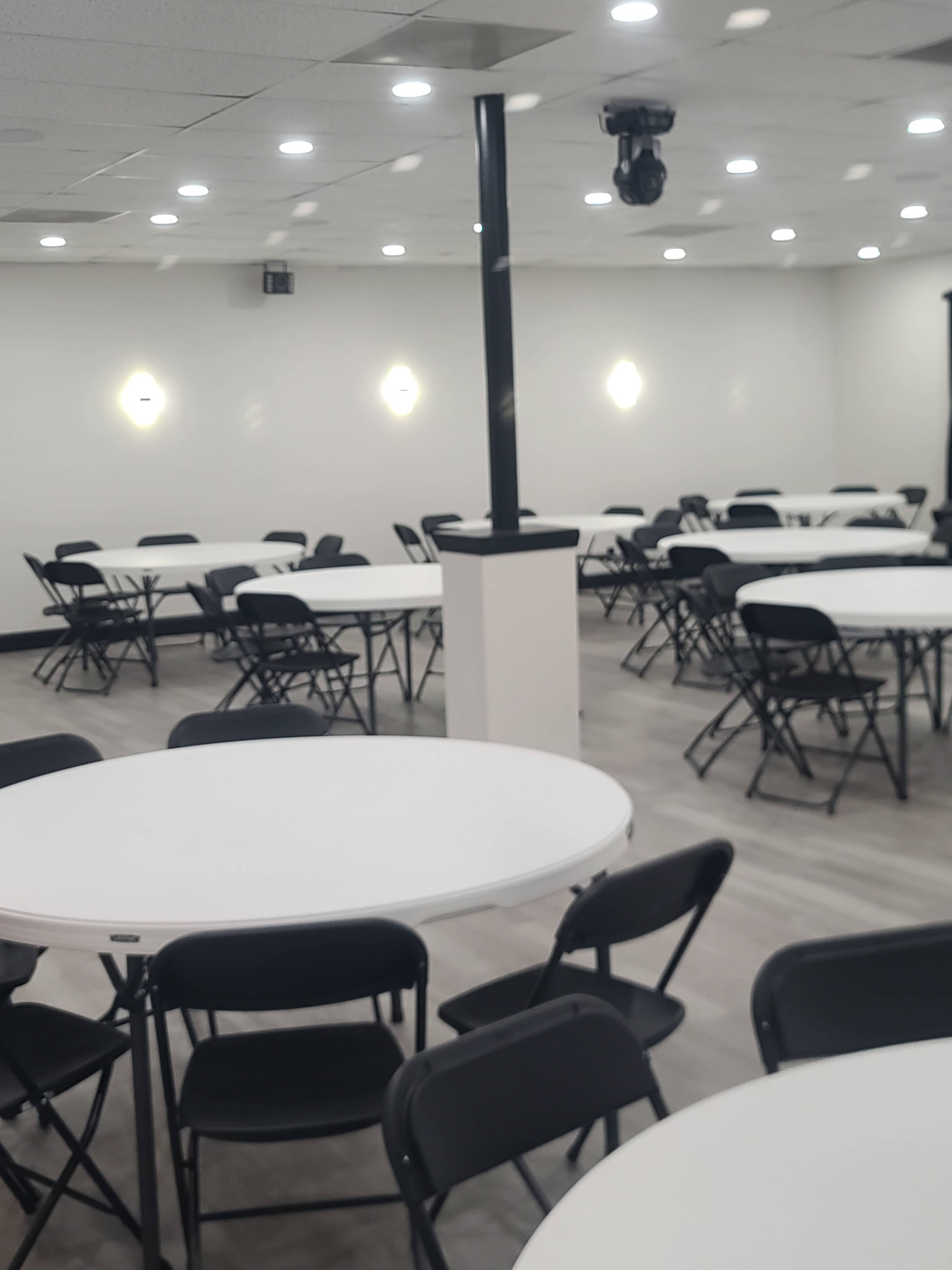 The image shows a well-lit room with multiple round tables and black folding chairs arranged neatly.