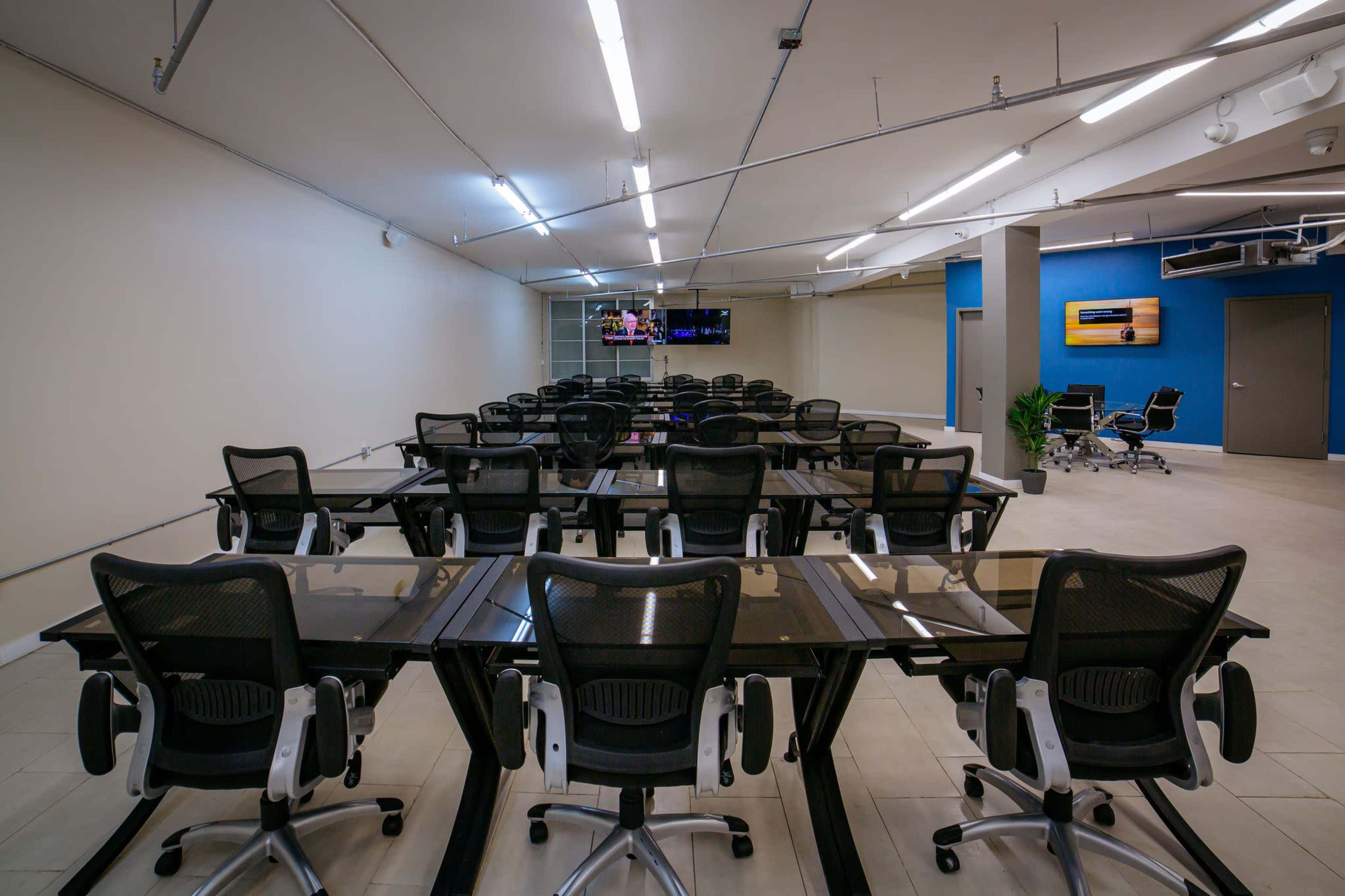 The image shows a spacious office with multiple desks and ergonomic chairs arranged in rows, featuring modern lighting and a blue accent wall.