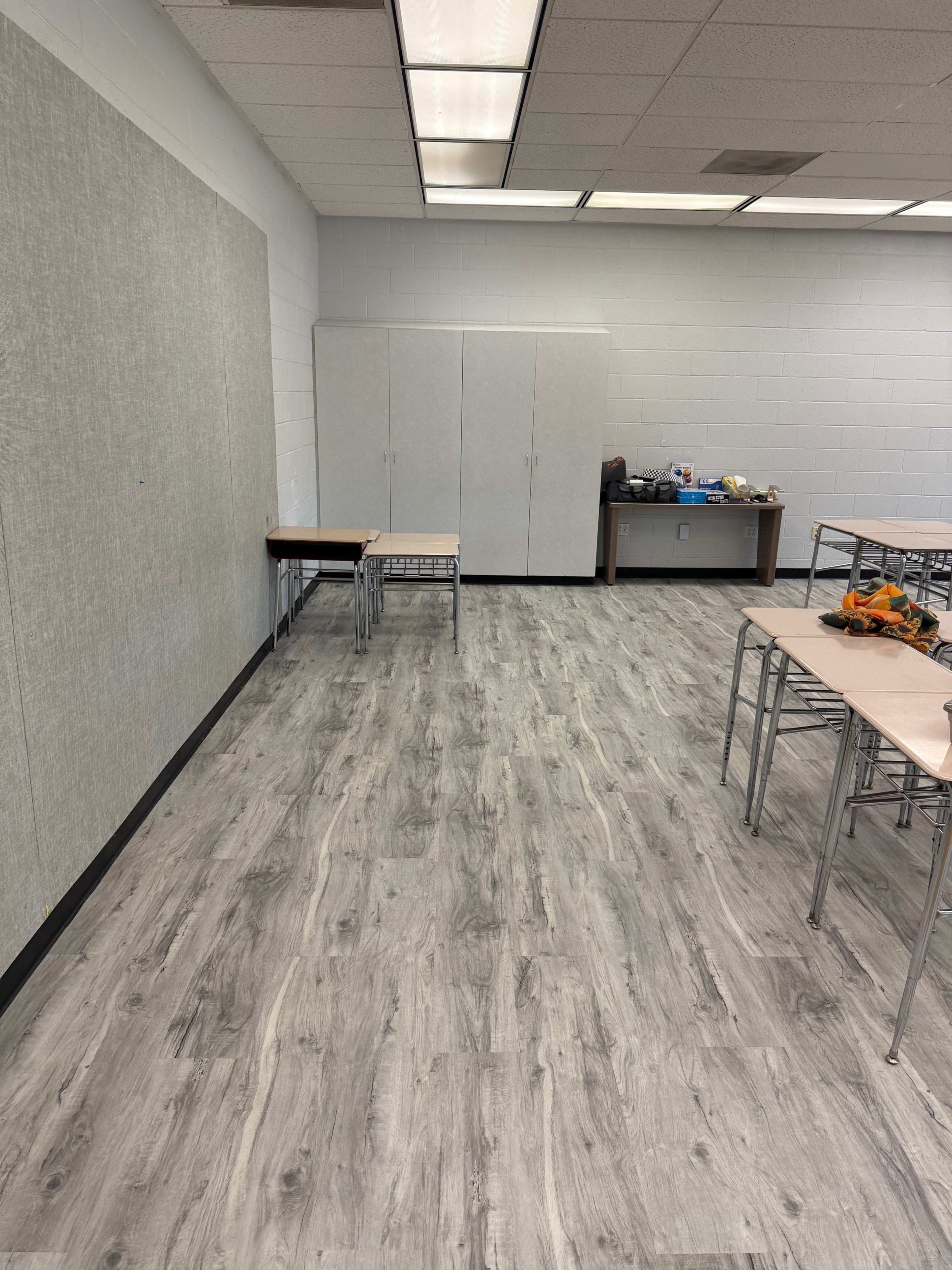 The image shows a sparsely furnished classroom with gray flooring, a whiteboard on one wall, and several empty tables arranged around the space.