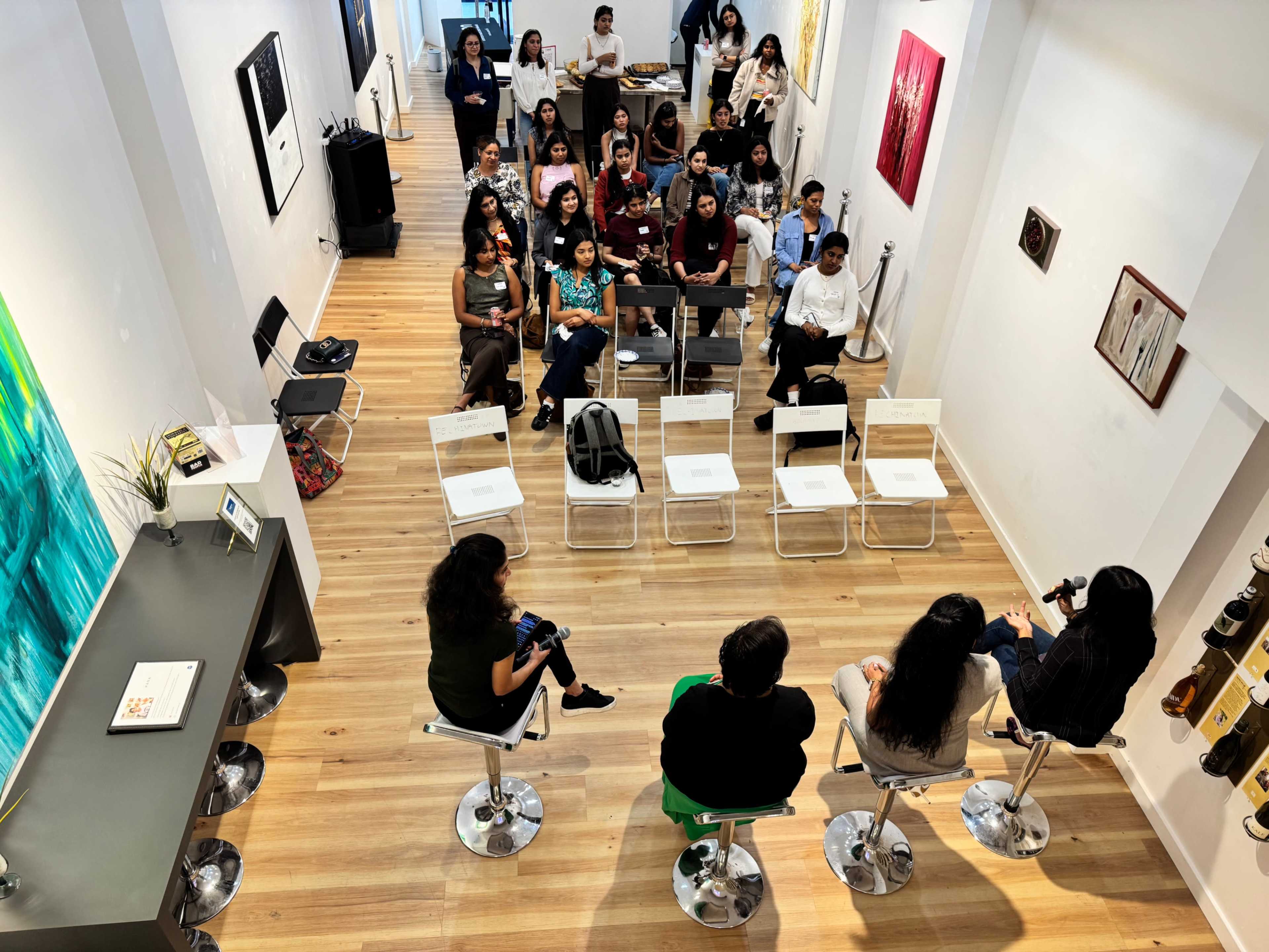 A diverse group of people is seated in an art gallery, facing a panel discussion taking place on a small stage at the front.