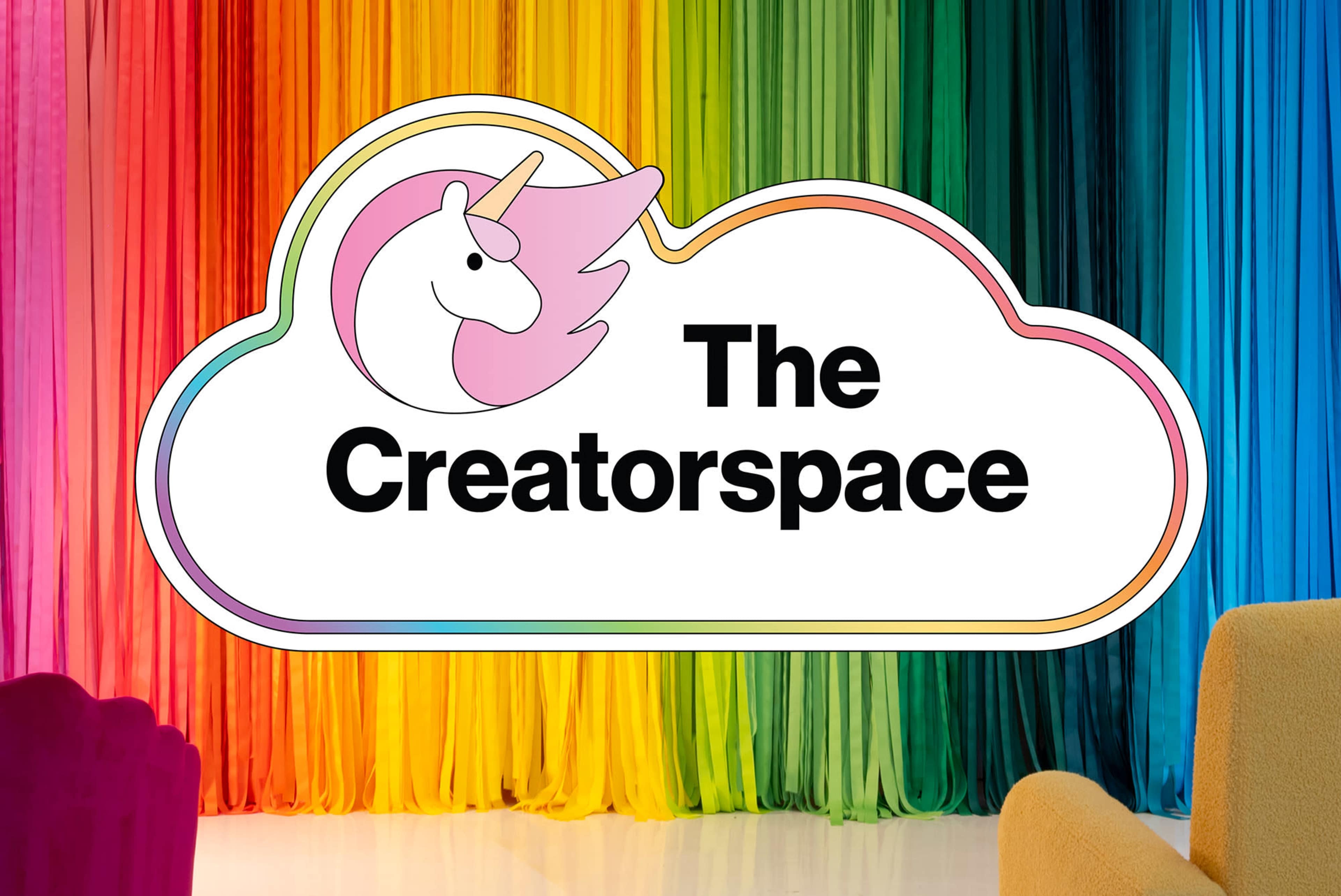 The image features a colorful backdrop of draped fabric in rainbow hues, with a sign display reading "The Creatorspace" alongside a unicorn graphic.