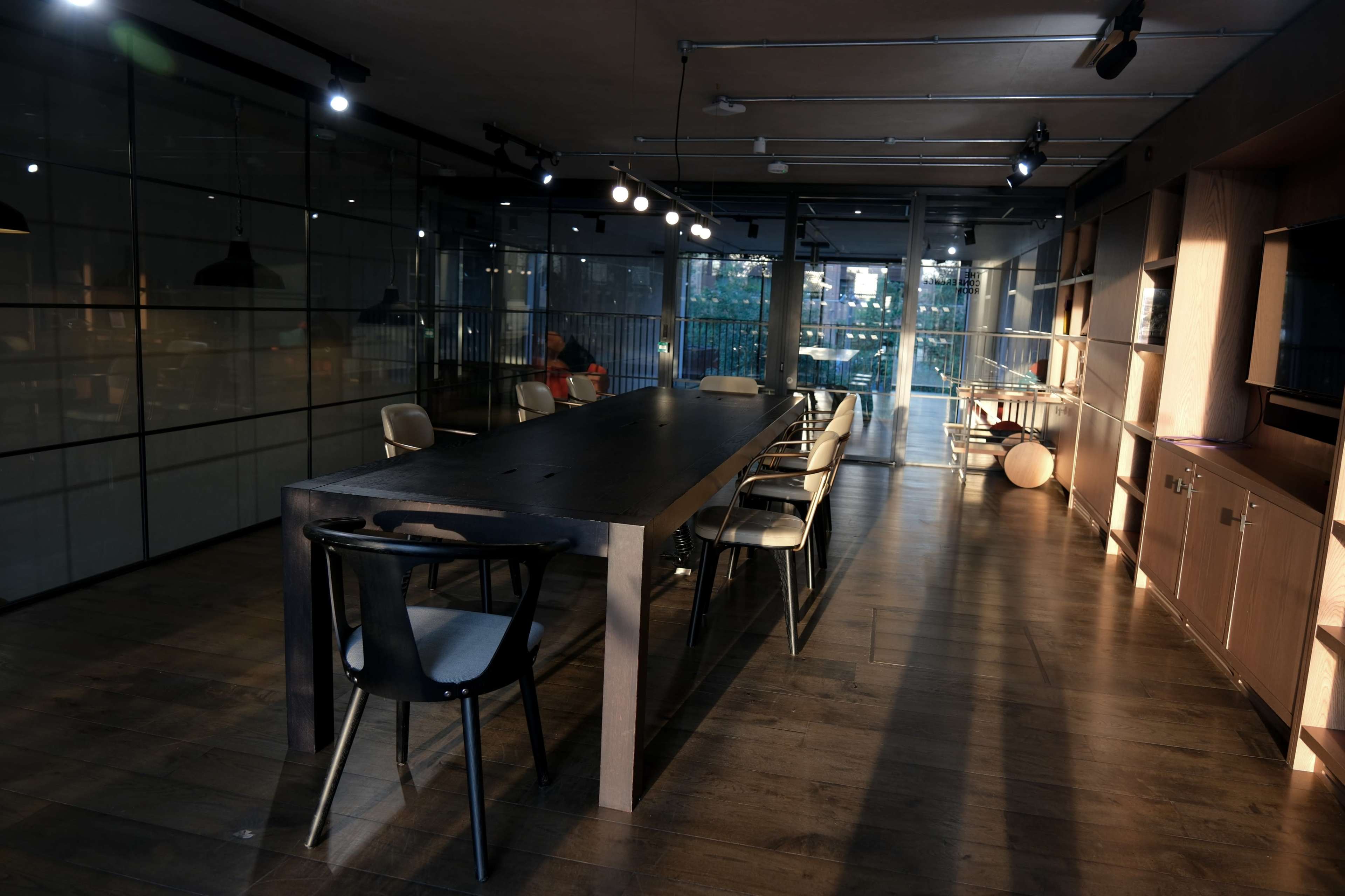 A modern, well-lit dining area with a long wooden table surrounded by chairs, and large windows providing natural light.