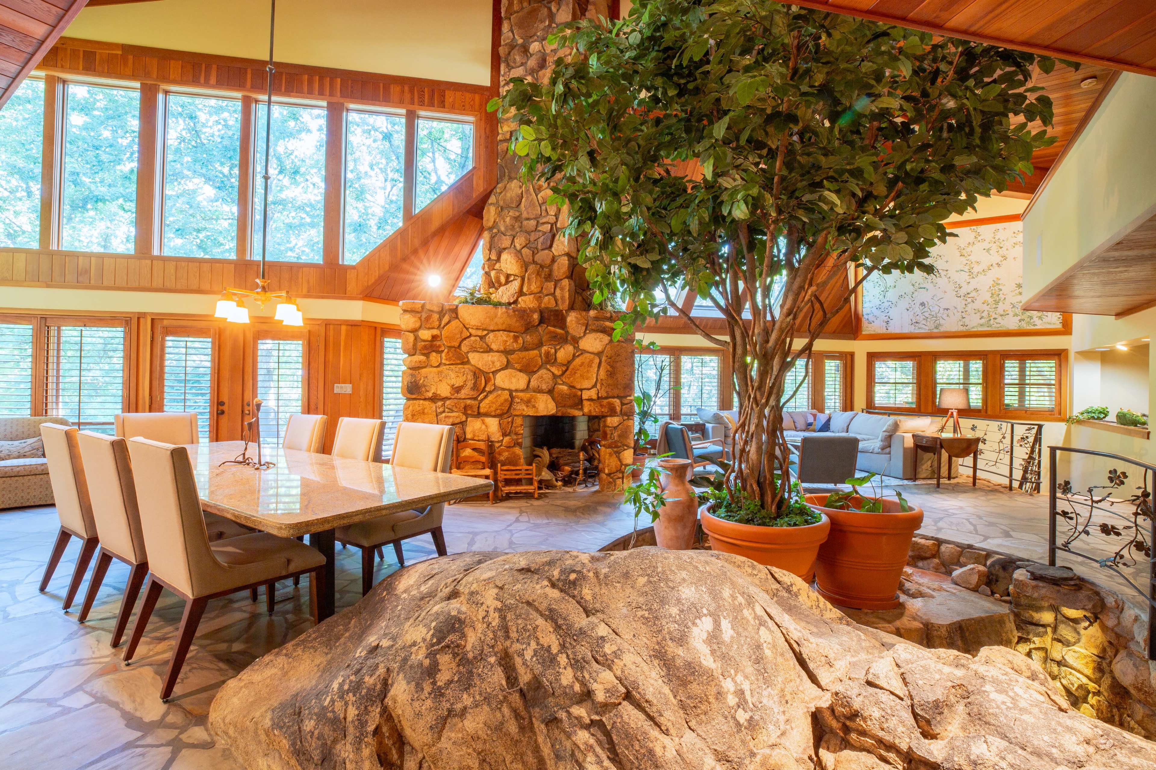 A spacious, rustic interior of a living area featuring a large stone fireplace, wooden beams, a dining table set for eight, and a decorative tree next to a large rock formation.