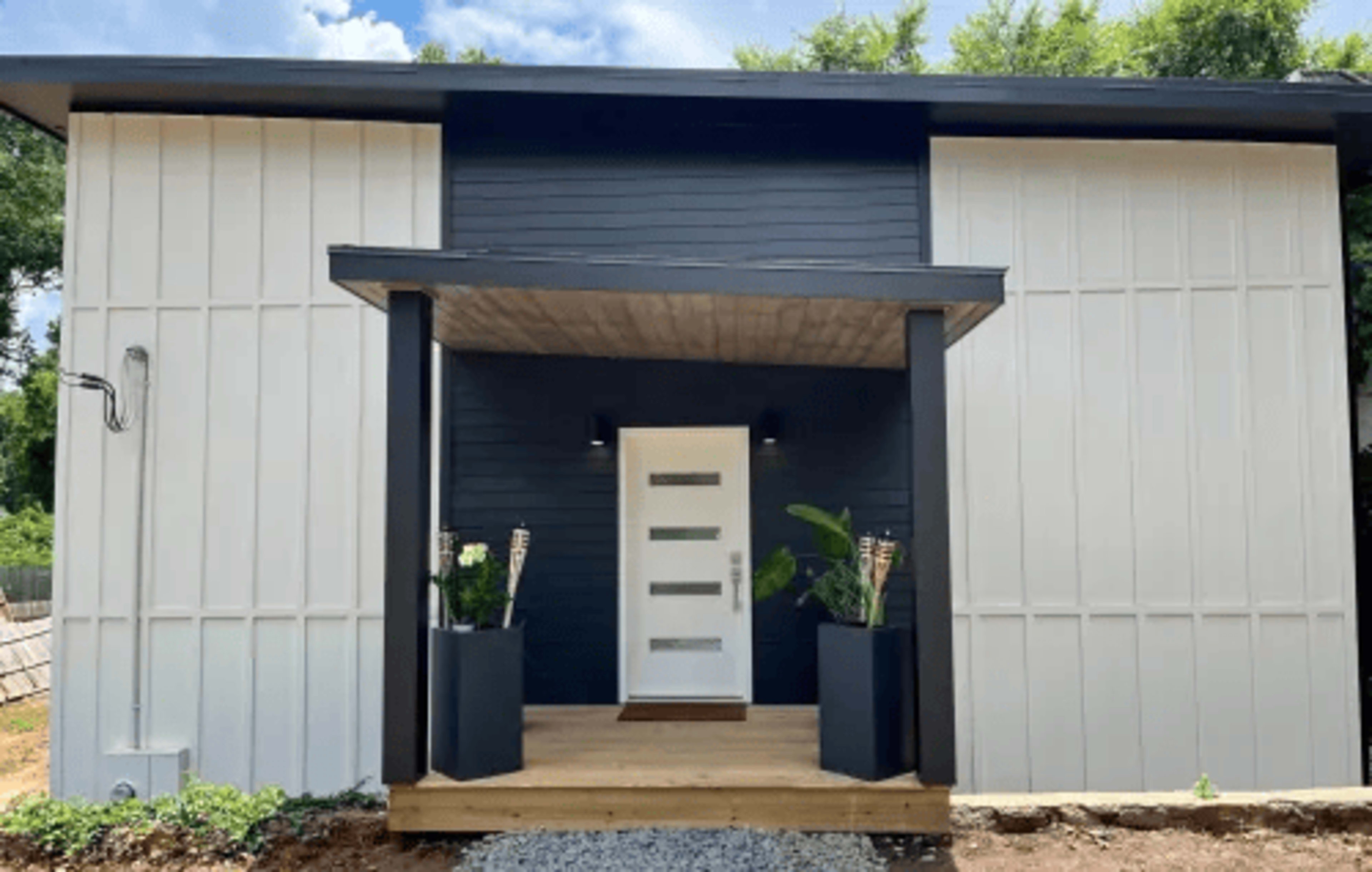 A modern house with a symmetrical facade, featuring a central white door and large planters on either side, set against a backdrop of contrasting white and dark blue siding.