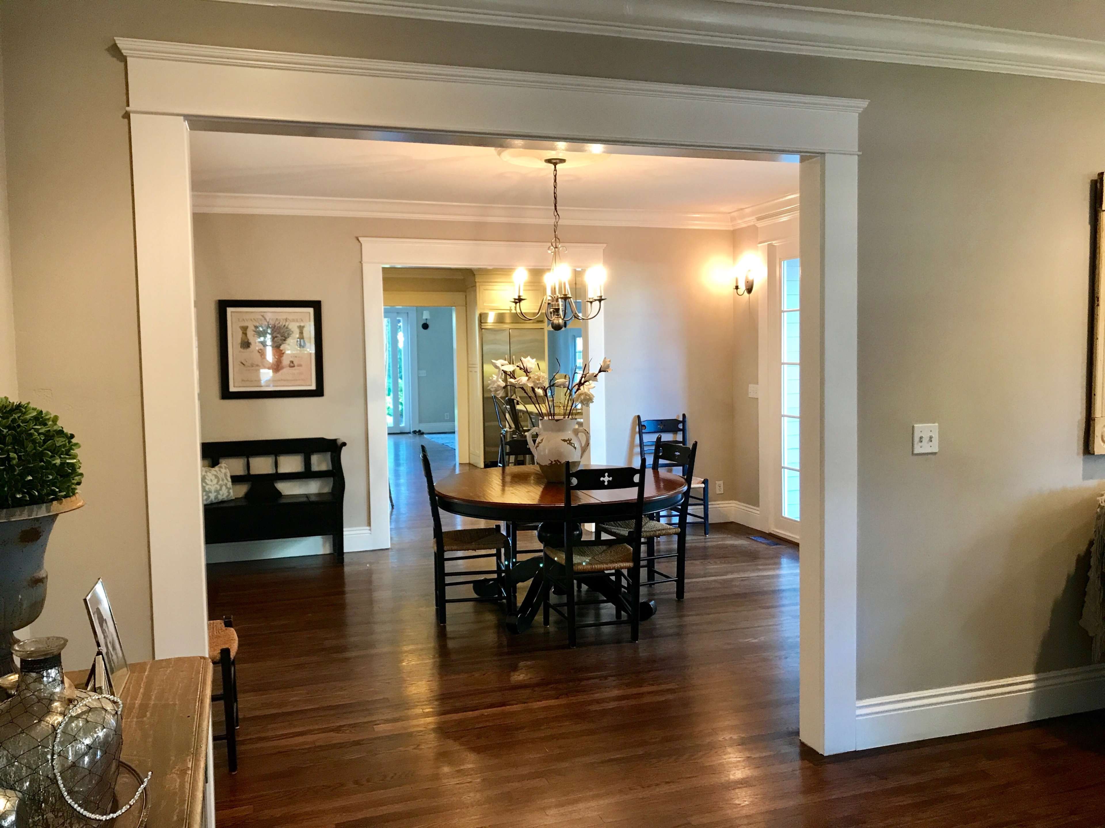 The image shows a dining area with a round wooden table and black chairs, framed by an open archway leading to another room.