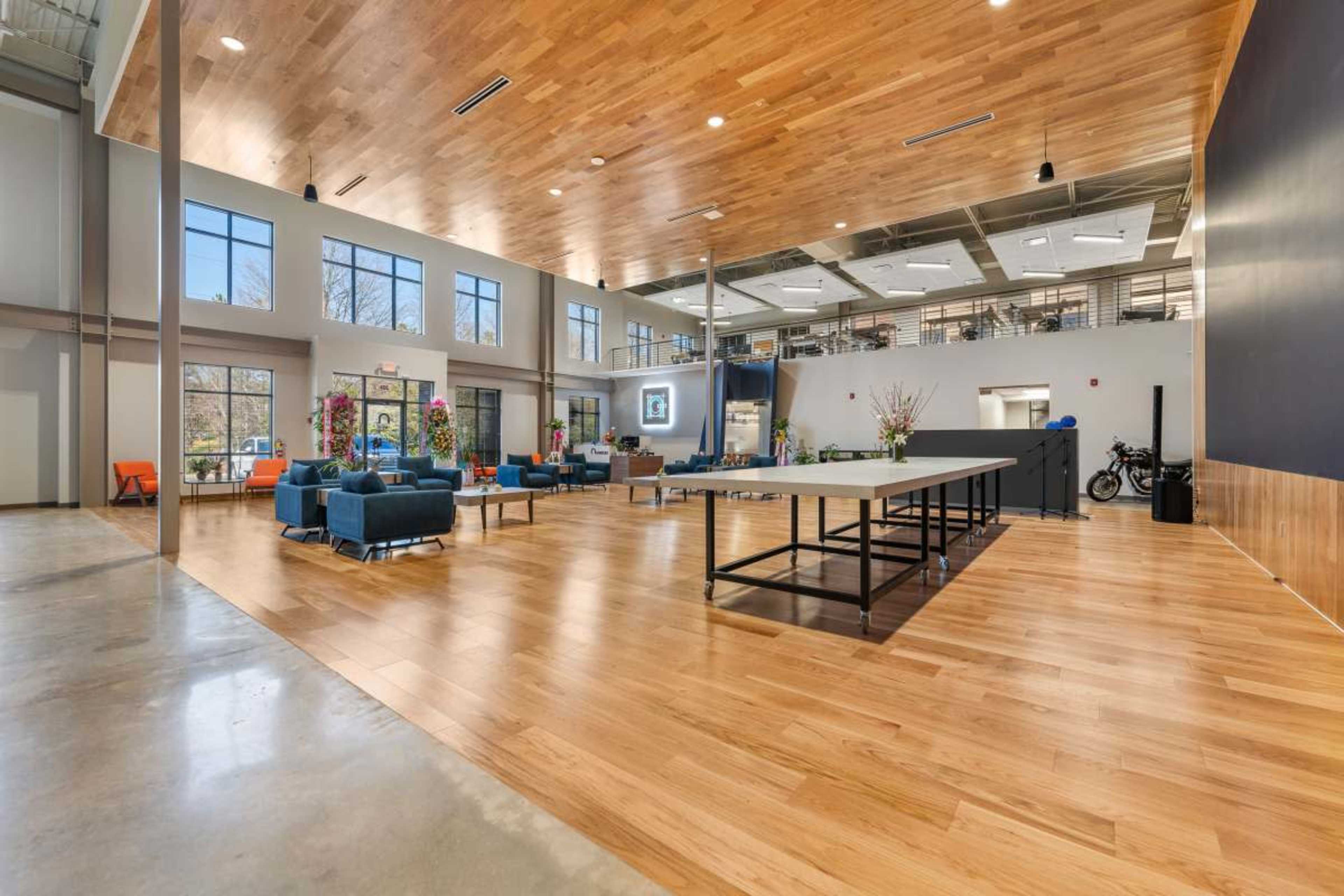 The image shows a spacious, modern interior of an office area featuring large windows, wooden flooring, and a mix of seating arrangements.