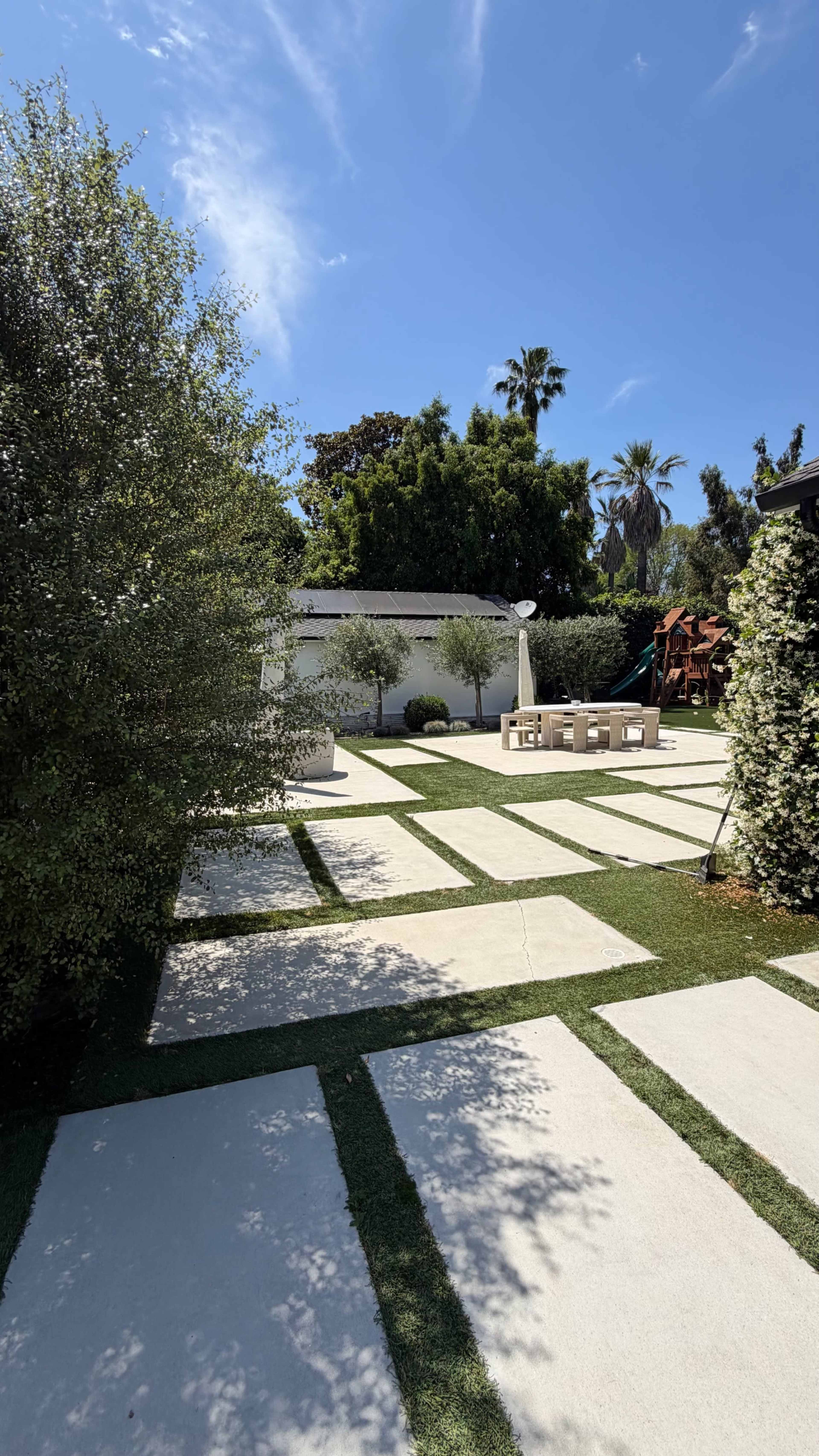 The image shows a neat backyard layout with rectangular concrete pathways, manicured grass areas, and greenery including trees and shrubs under a clear blue sky.