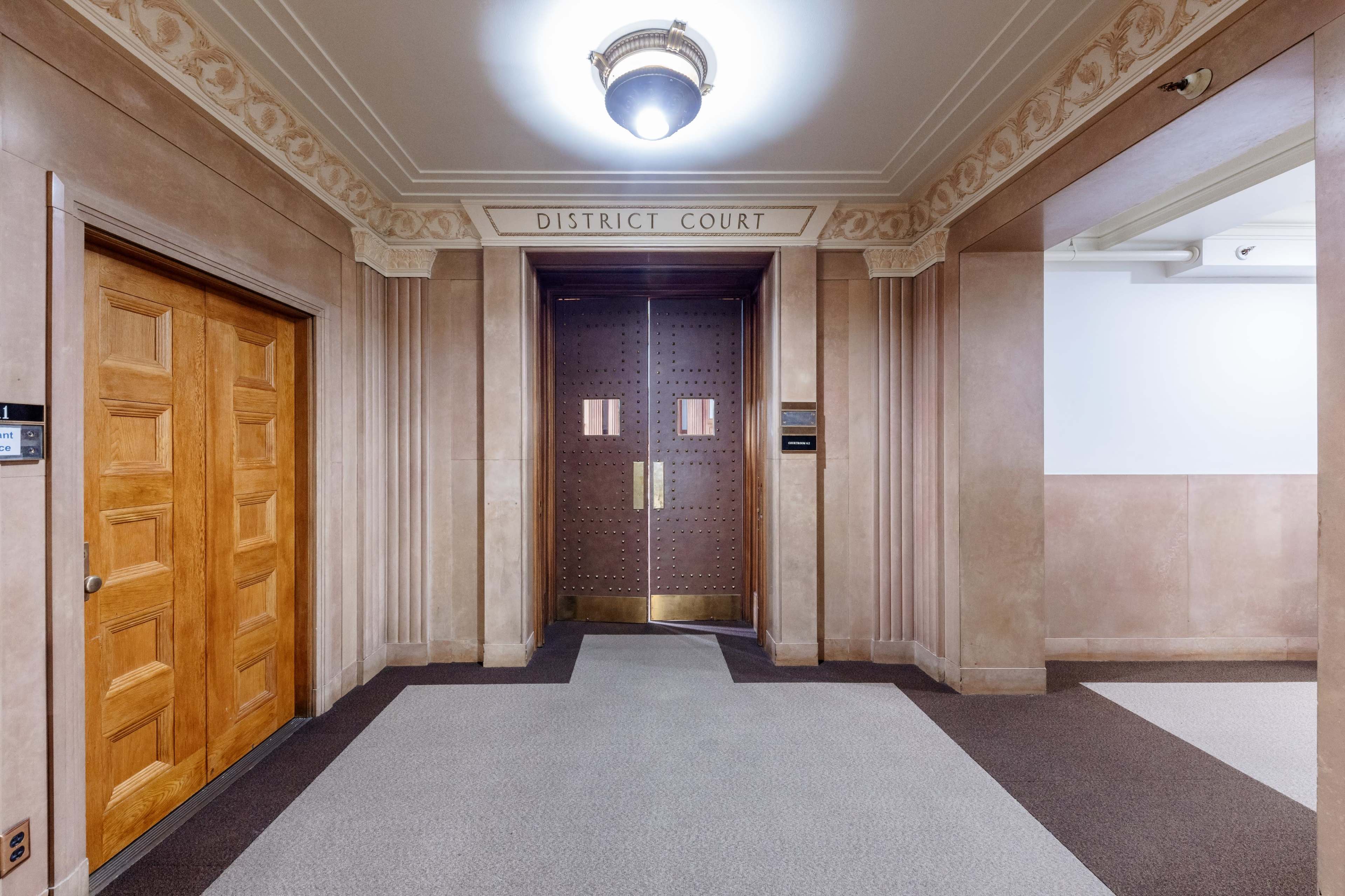 The image shows the entrance to a district court with double wooden doors and a decorative ceiling light in a corridor with patterned carpet.