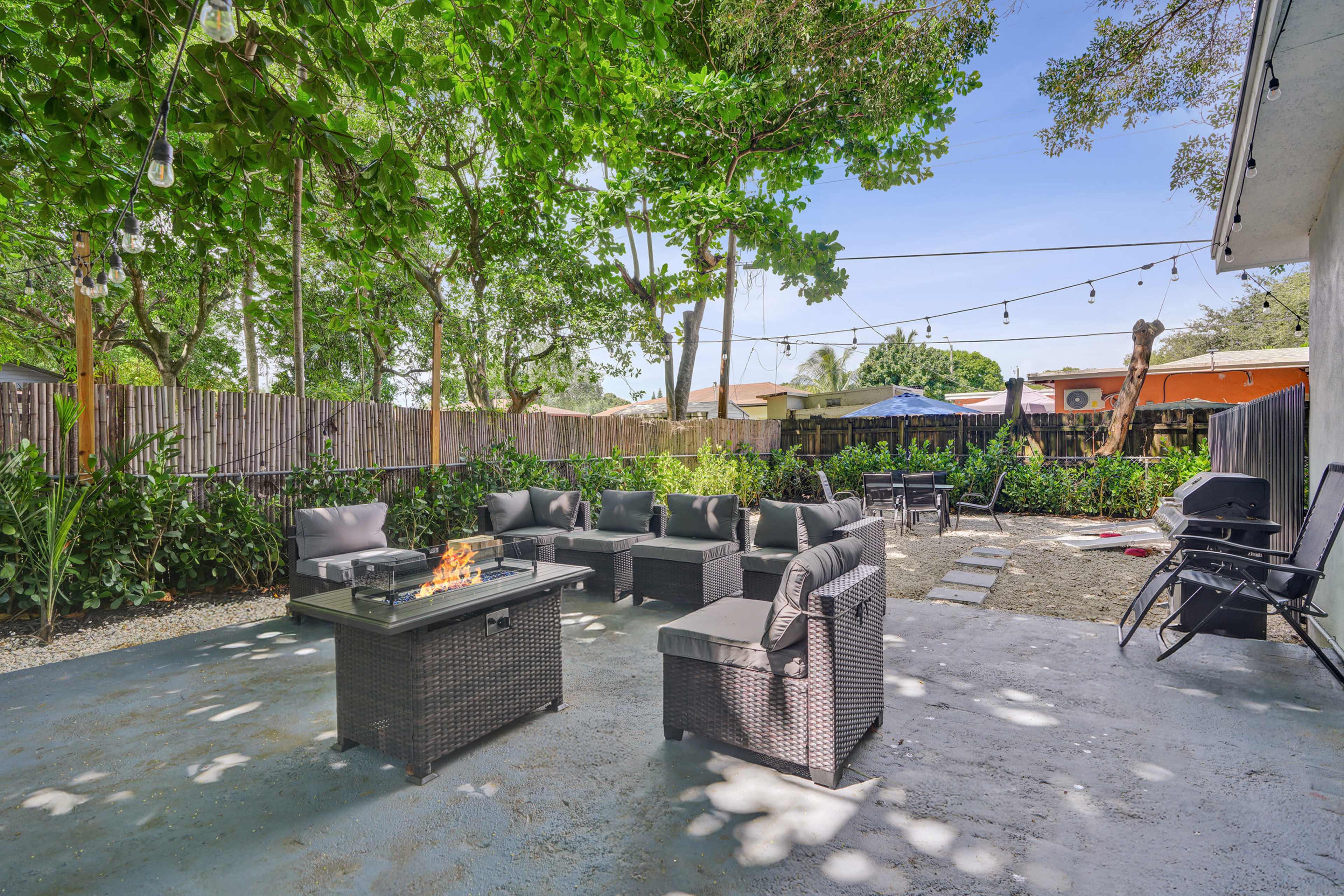The image shows a spacious outdoor backyard area featuring a fire pit surrounded by comfortable seating, with lush green trees and garden plants in the background.