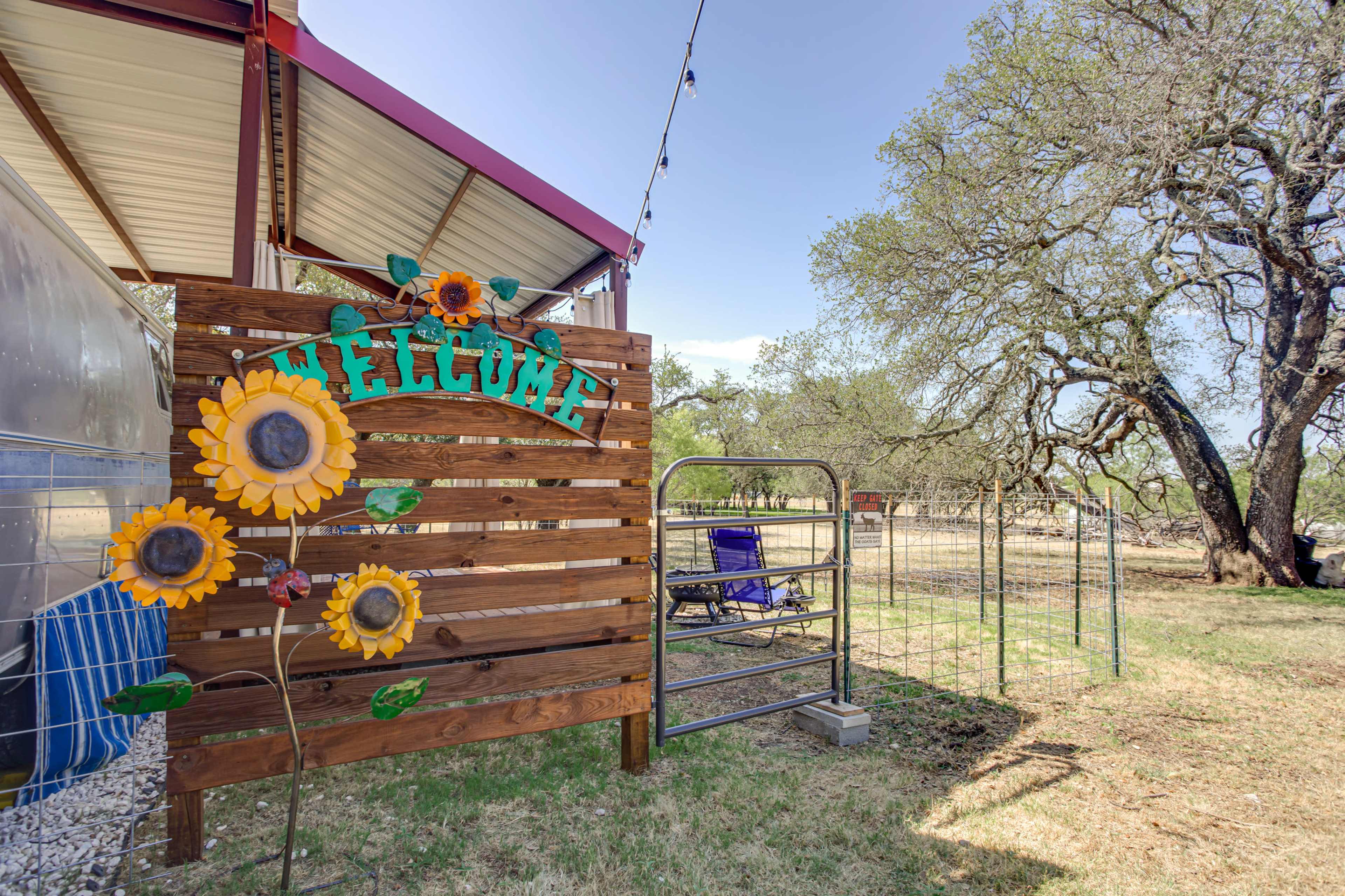 A wooden welcome sign adorned with sunflowers stands near a fenced area and an open space with an oak tree in a rural setting.