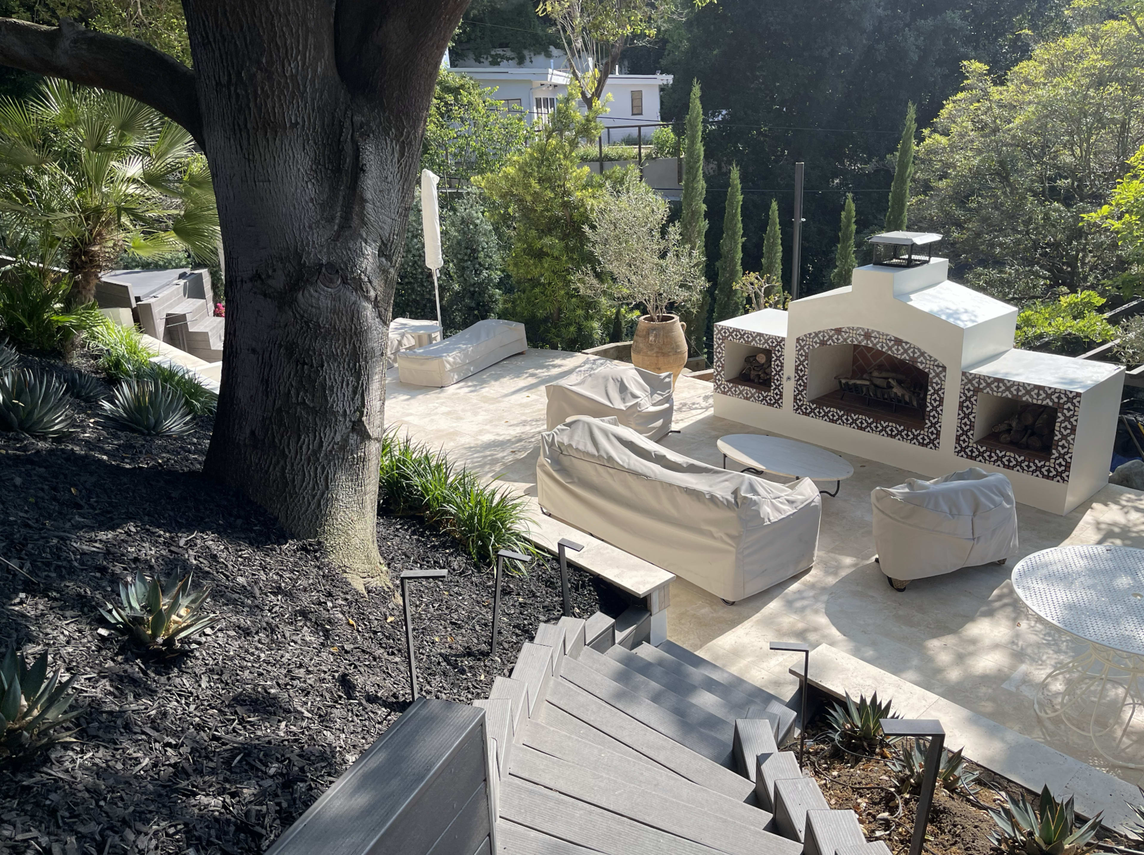 The image shows a landscaped outdoor area with a stone patio, seating arrangements, and a fireplace surrounded by greenery.