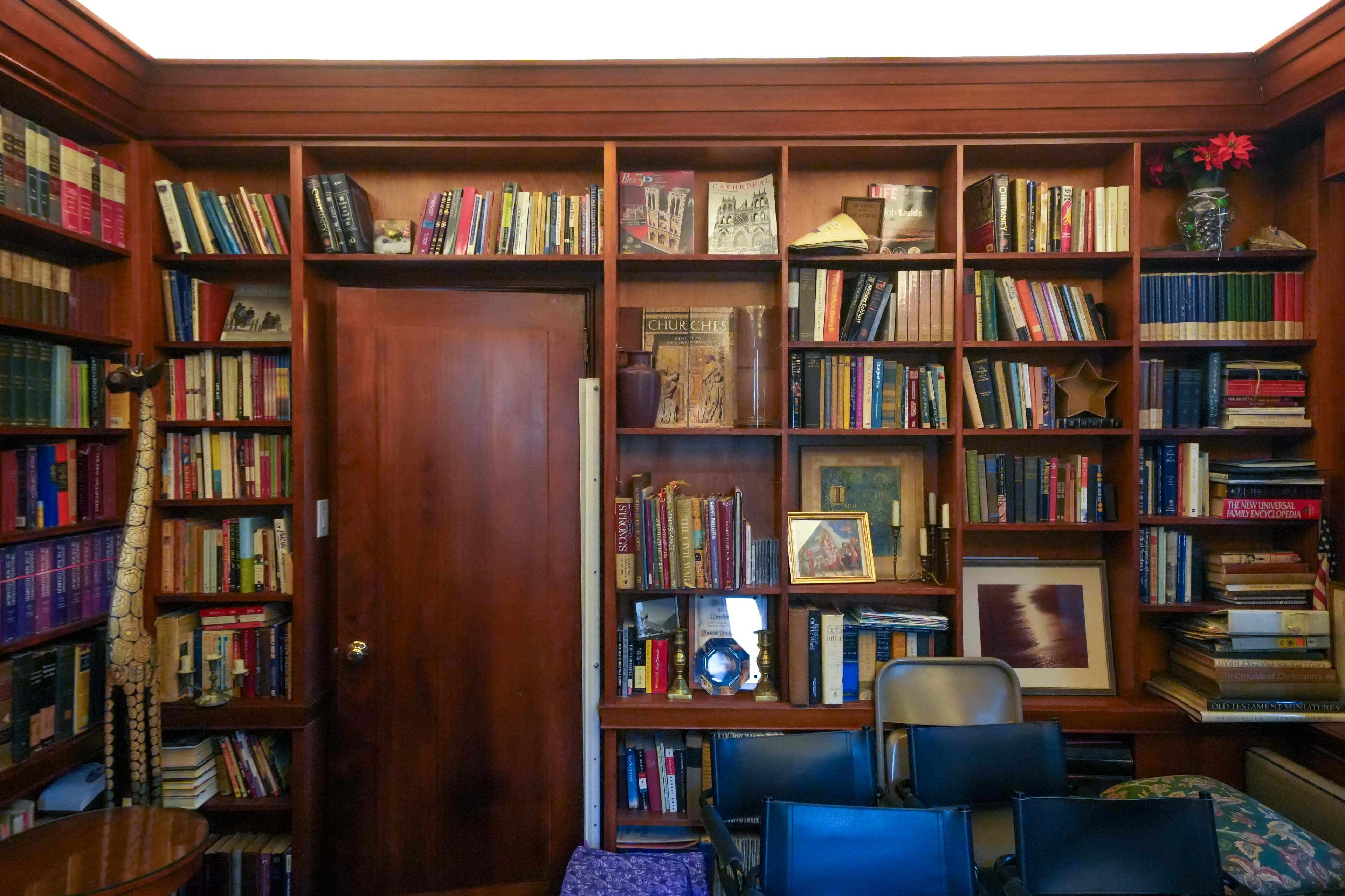 A wooden bookshelf filled with various books and decorative items, adjacent to a closed door and a stack of chairs in a cozy room.